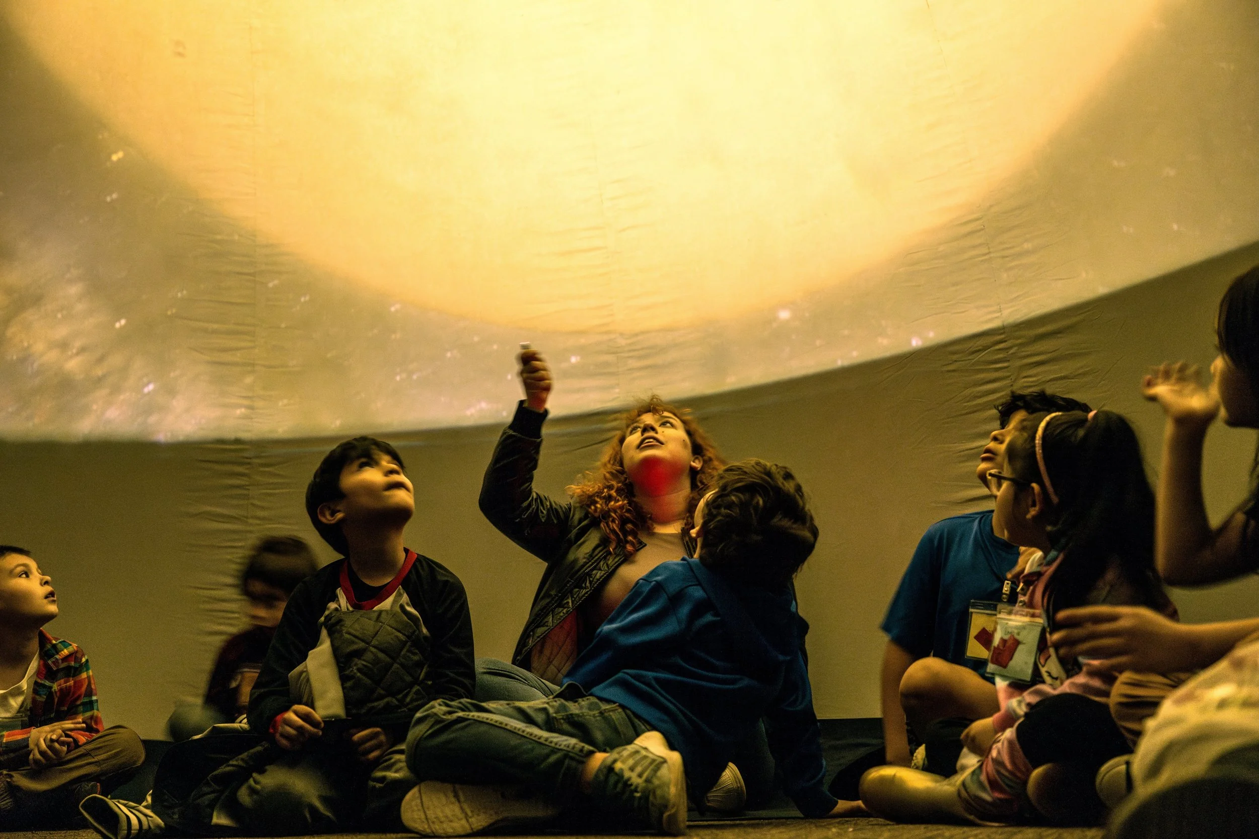 A Solstice Outreach planetarium presenter sits surrounded by young elementary-age children holding a laser pointer pointed up at a projection of the sun on the planetarium dome.