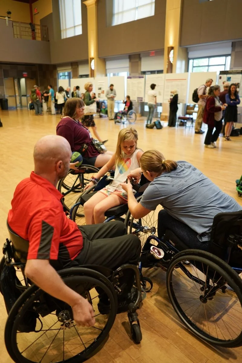 A young girl sits in an active wheelchair as another wheelchair user adjusts the straps on it for her. A third wheelchair user watches. The users are in a gymnasium, with conference posters in the background.