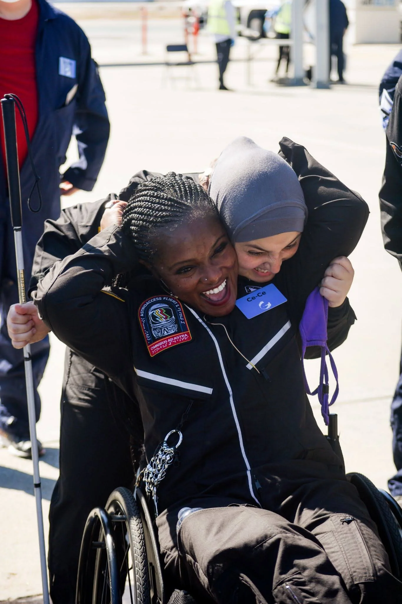 AstroAccess Ambassador Ce-Ce Mazyck, a black woman with braids wearing a black flight suit, sits in her wheelchair laughing as she hugs AstroAccess Mona Minkara, a woman wearing a hijab and carrying a white cane, who is hugging Ce-Ce from behind.