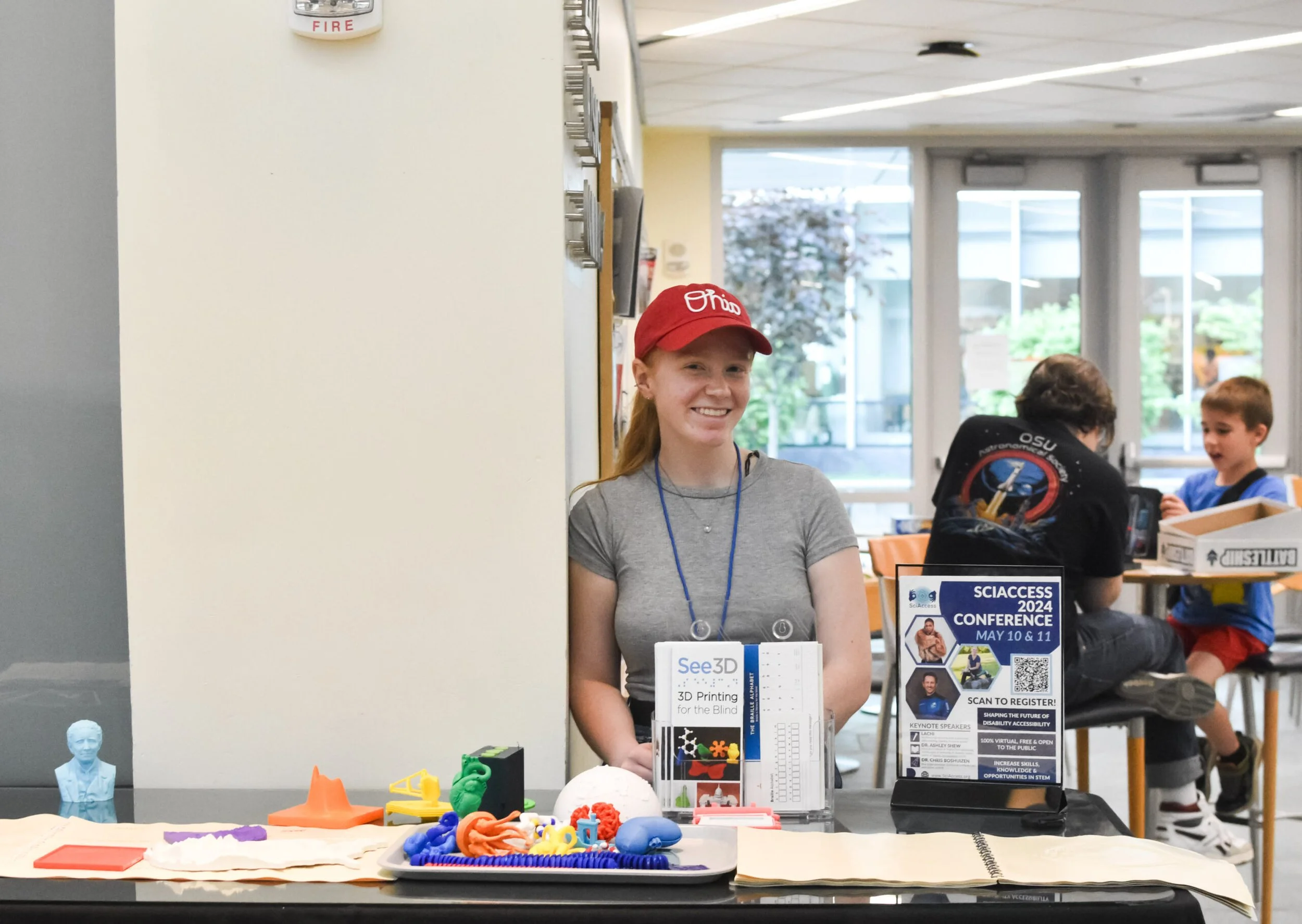 A SciAccess volunteer, a young white woman wearing a red script Ohio hat, stands behind a table displaying 3D models and materials advertising the SciAccess 2024 conference.