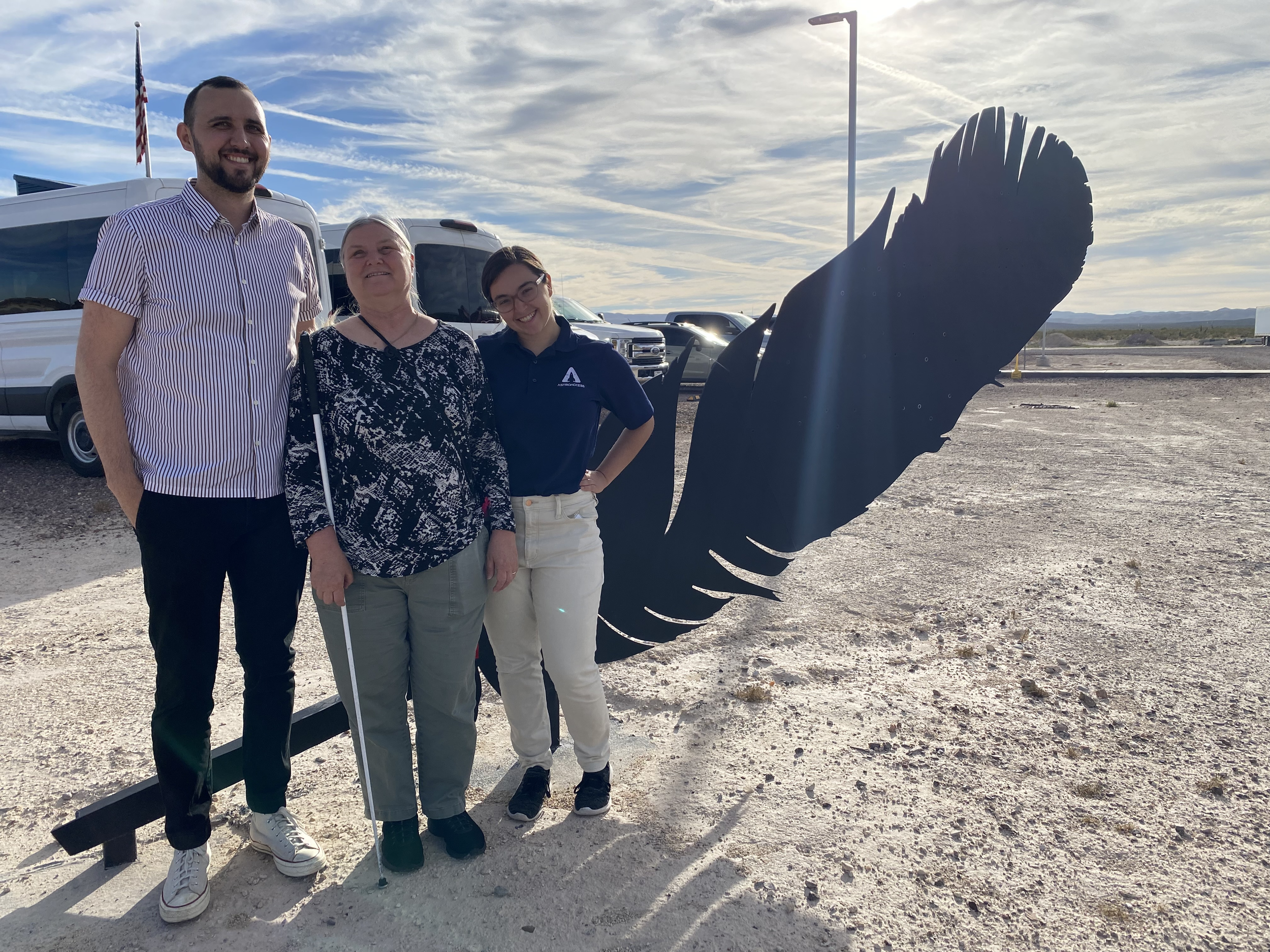 Three AstroAccess team members smile in front of a statue of the Blue Origin feather logo. One person wears a blue AstroAccess polo, and one carries a white cane.