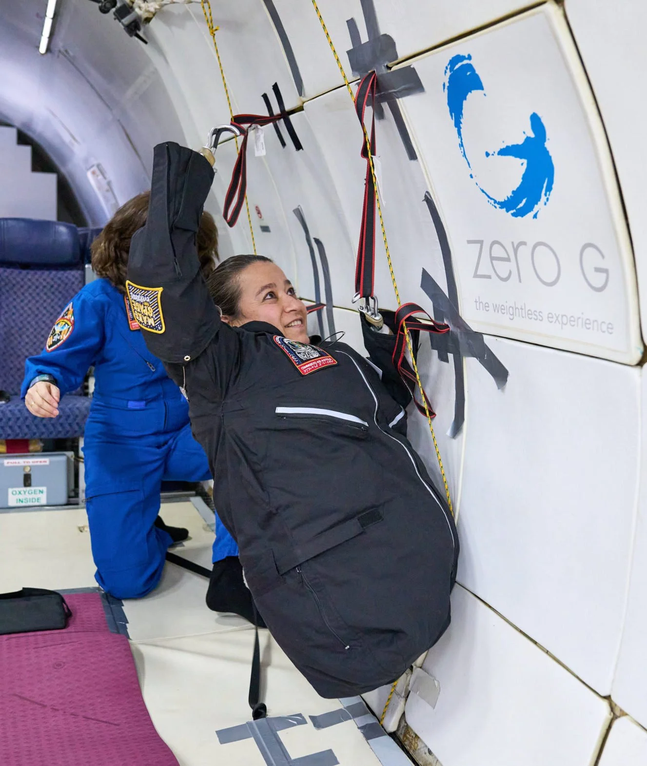 AstroAccess Ambassador Dana Bolles onboard a zero-gravity flight, using her hook arm prosthetics to pull herself up the wall of the cabin using straps attached to the wall.