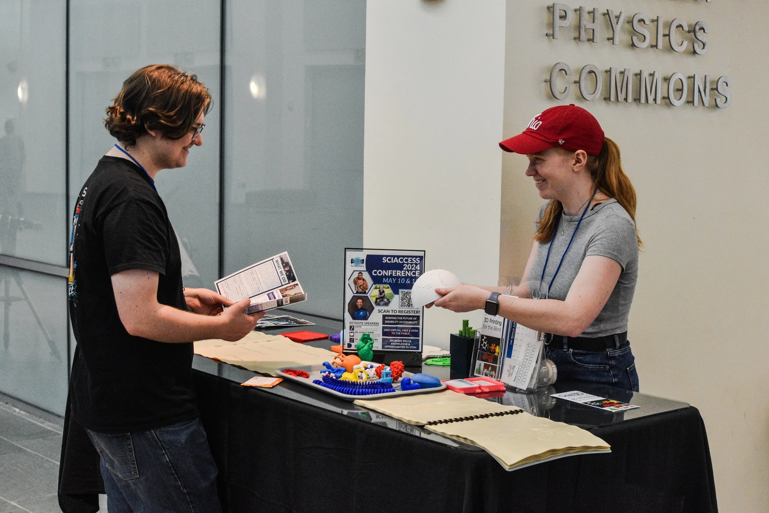 A SciAccess volunteer, a young white woman wearing a red hat, holds out a white domed 3D model to a man reading a pamphlet in front of a table of 3D models and materials about SciAccess.