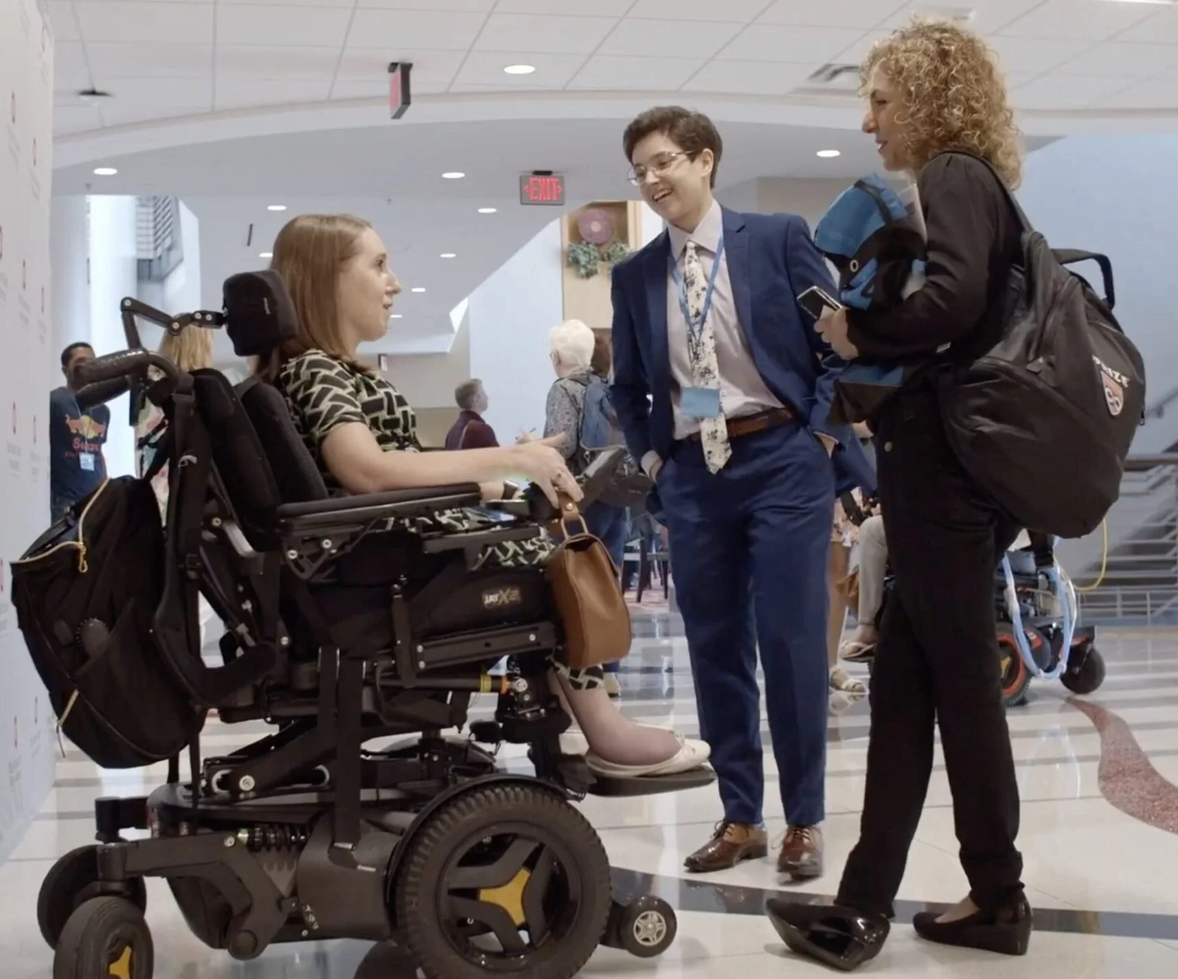 SciAccess Executive Director Anna Voelker, a nonbinary person in a blue suit, speaking with Miss Wheelchair USA 2018 Heather Tomko, a white woman in an electric wheelchair, and Anousheh Ansari, an Iranian-American engineer and first Iranian astronaut