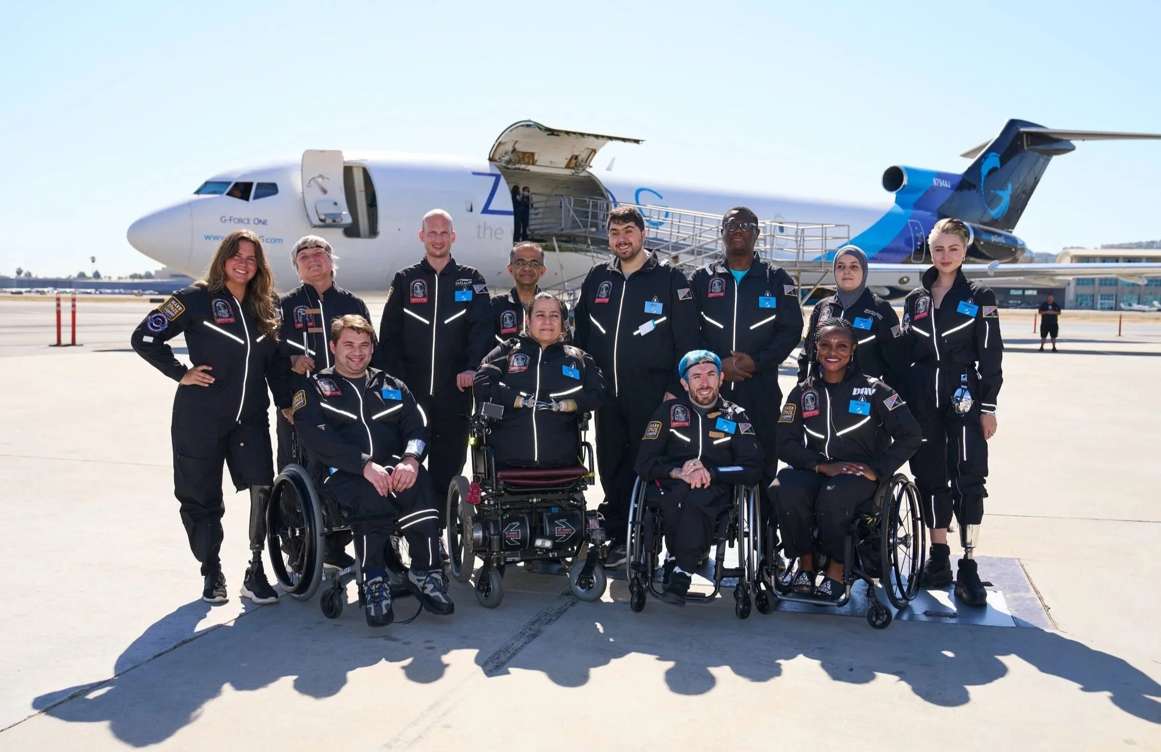 AstroAccess Ambassadors smile for a group photo on the tarmac in front of the Zero-G plane. Twelve people of varying ethnicities and genders are picture. Four people are wheelchair users, two have visible prosthetics, and two hold white canes.