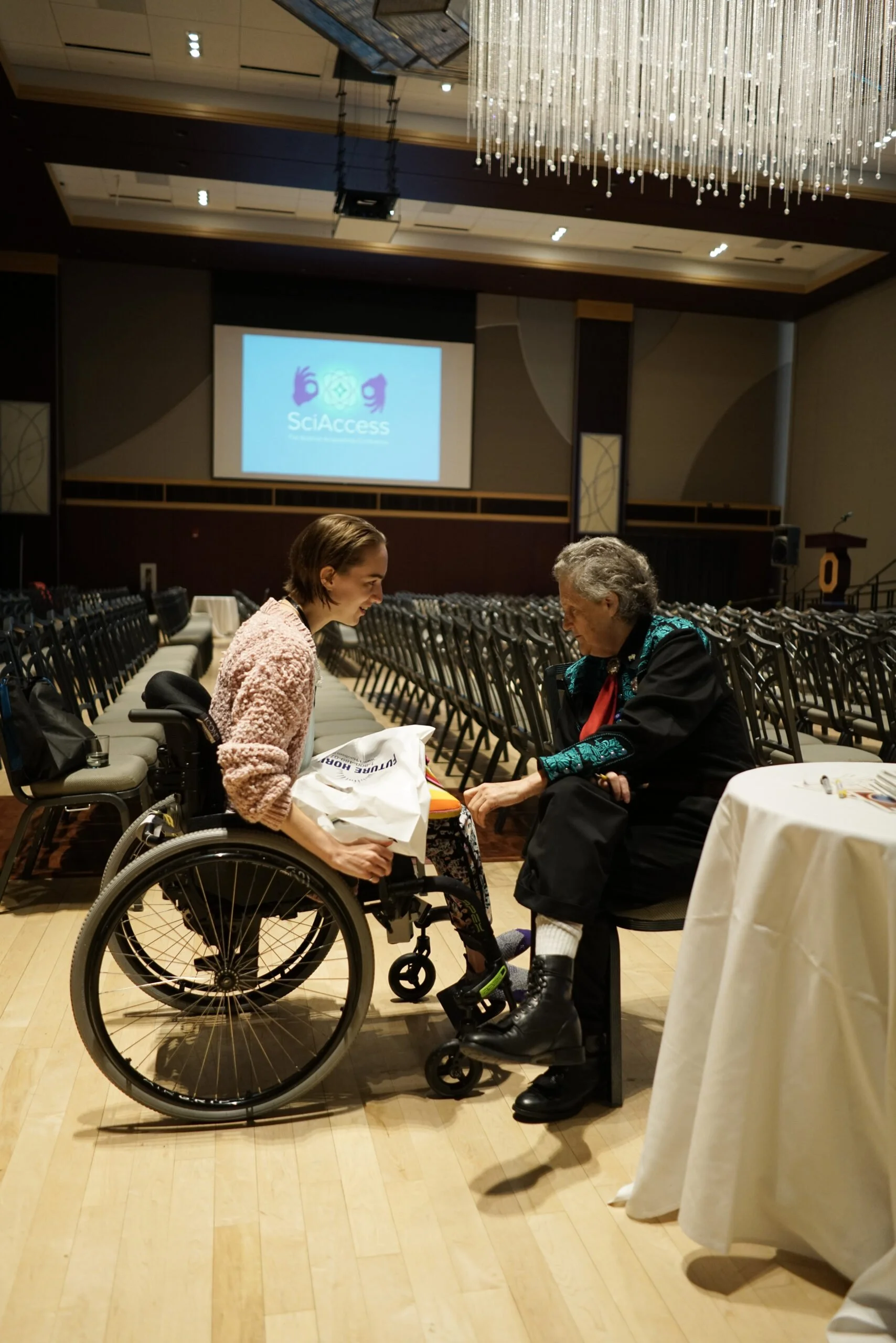 Biomedical engineer and wheelchair user Jen Schlegel speaks with renowned autism self-advocate Temple Grandin in an empty conference hall talk following Grandin's keynote presentation. The SciAccess logo is projected on a screen behind them.
