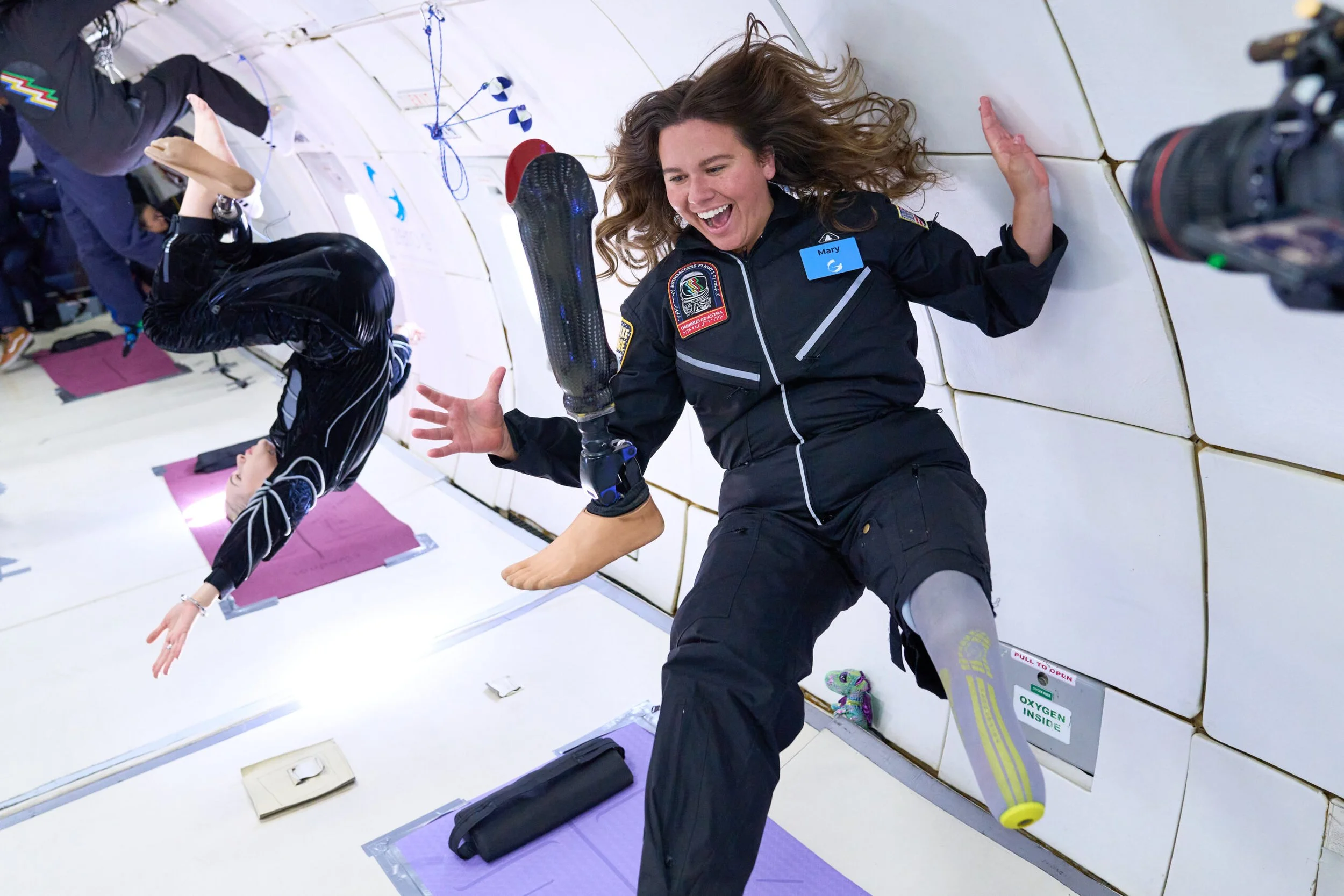 AstroAccess Ambassador Mary Cooper, a young white woman, floats in zero gravity. She has removed her prosthetic leg and is floating it in the air in front of her. She is wearing a black flight suit with patches and smiling broadly.