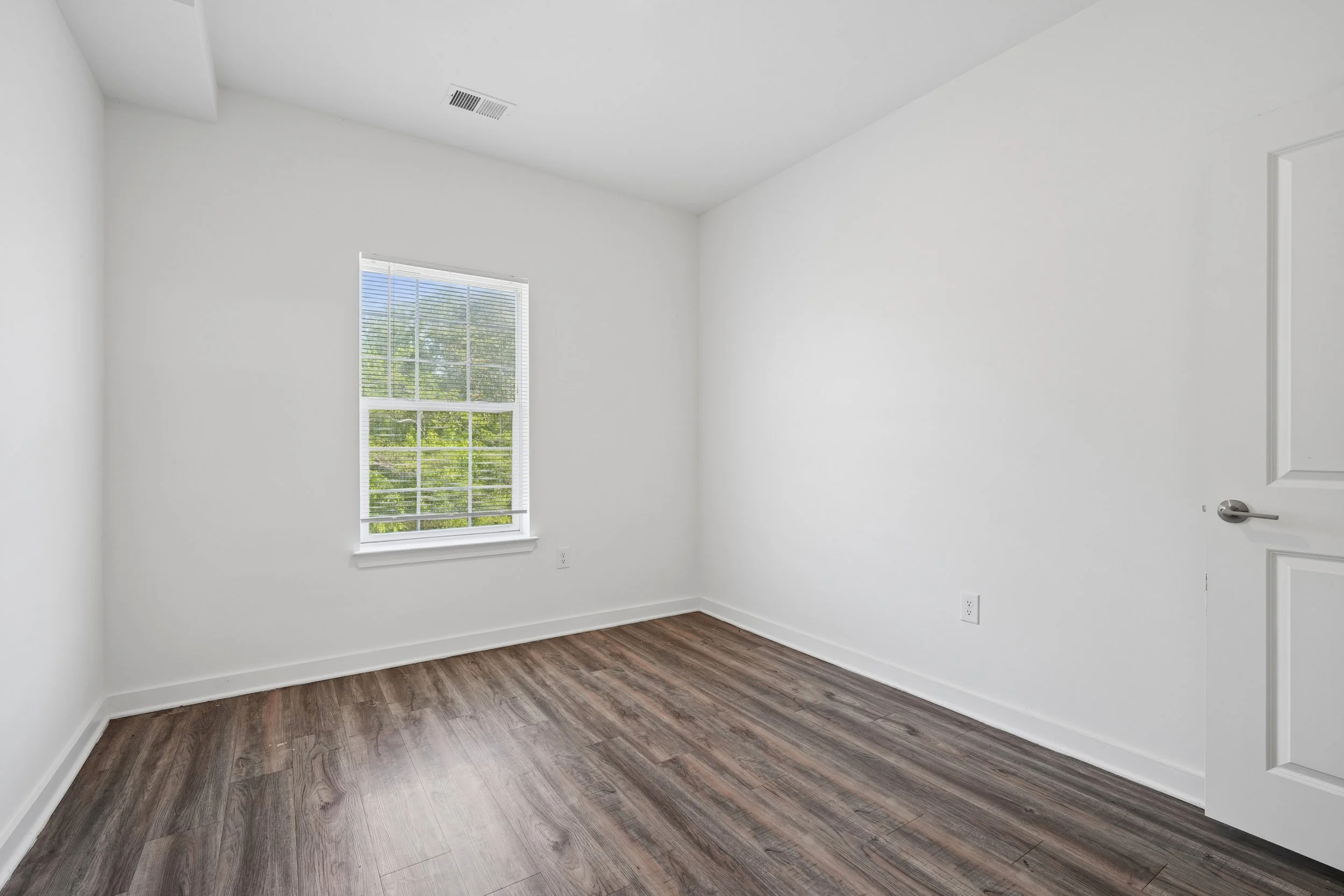 Empty room with white walls, a window with white blinds showing trees outside, wood laminate flooring, and a closed white door.