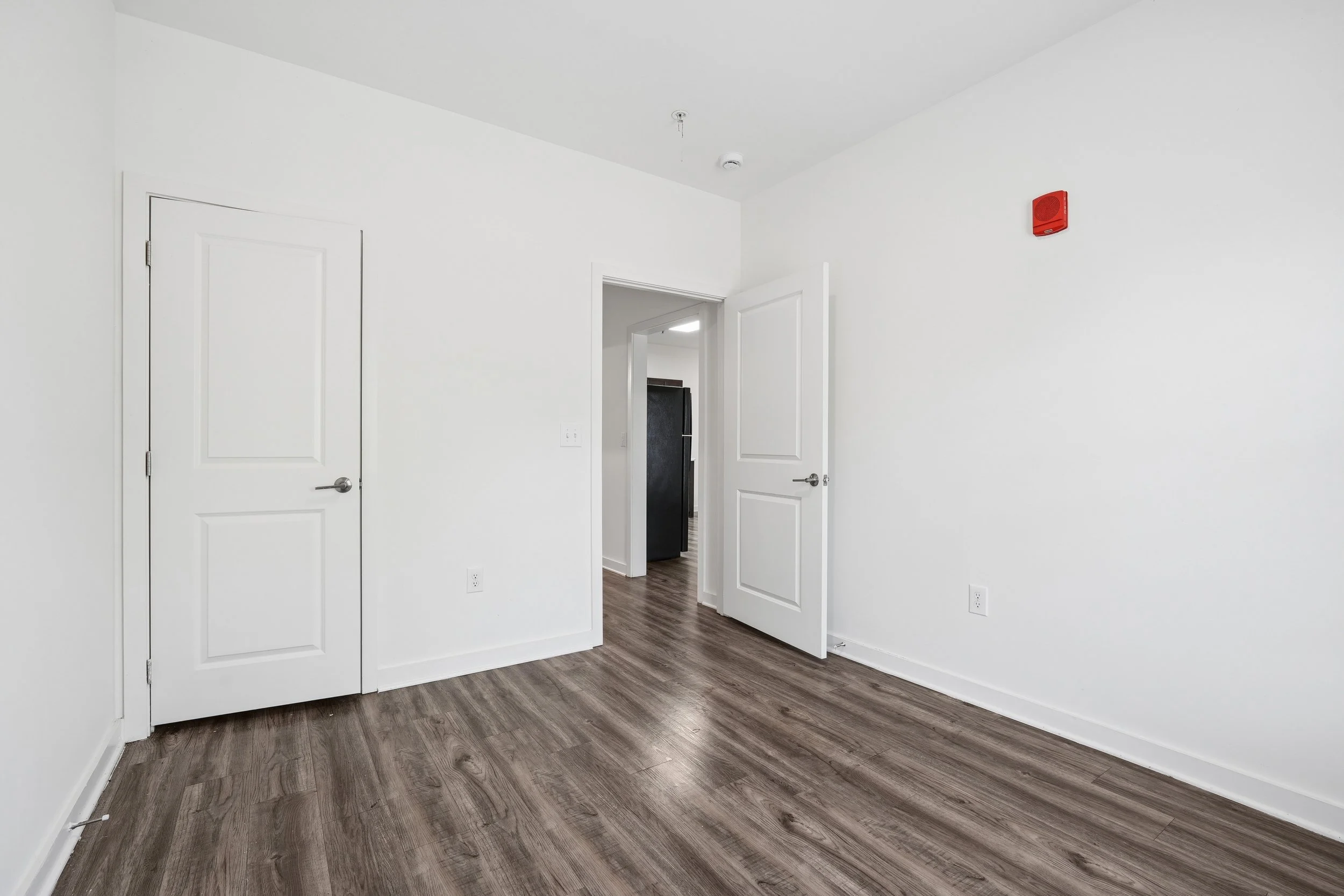 Empty room with white walls, wood flooring, closed and open doors, and a red fire alarm on the wall.