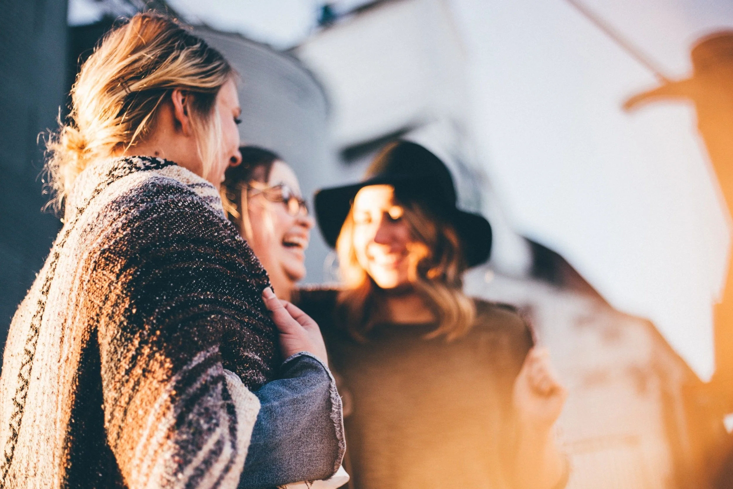 Four women with different hair colors and styles, wearing casual clothing, are standing close together, smiling, and appearing to be engaged in a friendly conversation.