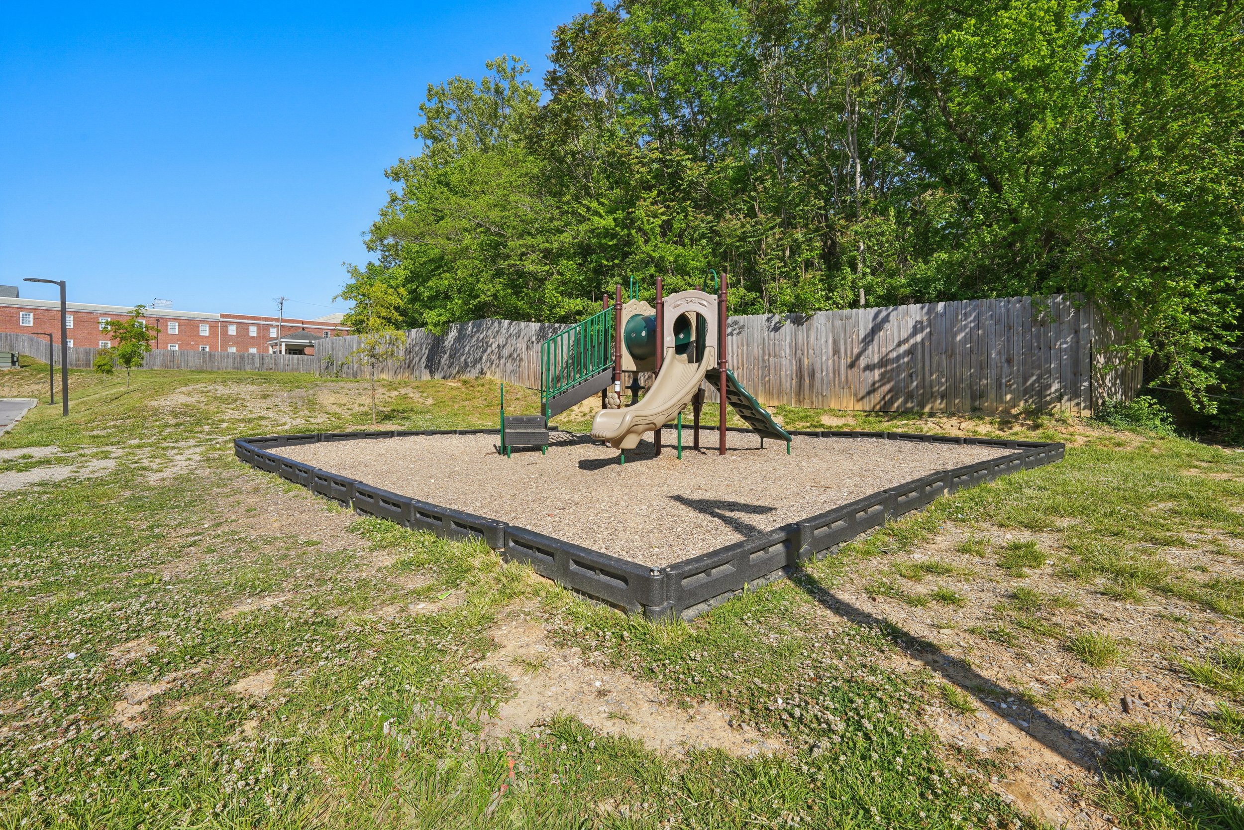 A small playground area with a slide and climbing structure, surrounded by a black border, situated on a grassy field with patches of dirt. There are trees and a wooden fence in the background, with a brick building visible in the distance under a cl