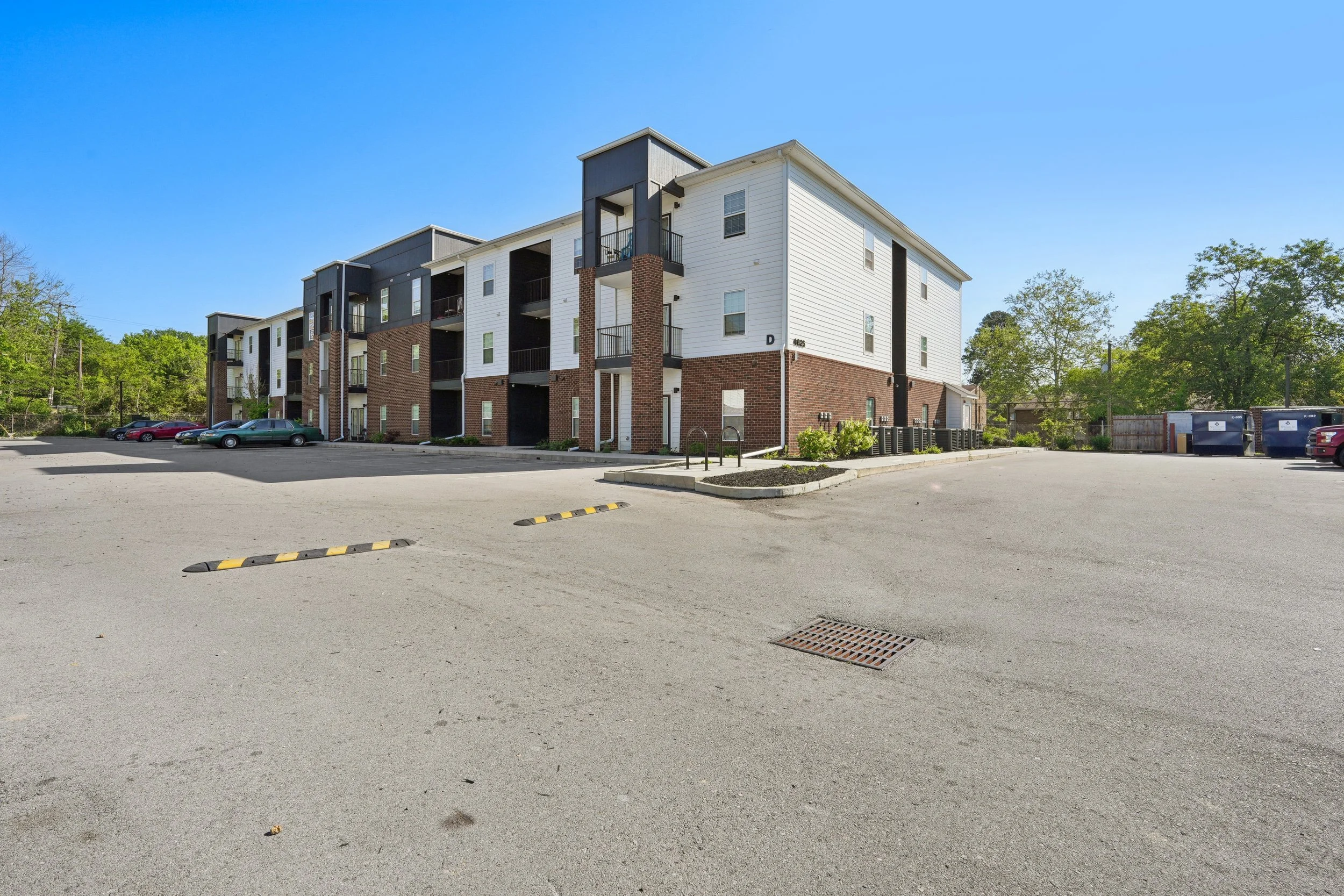 An empty parking lot in front of a modern multi-story residential building with trees in the background.