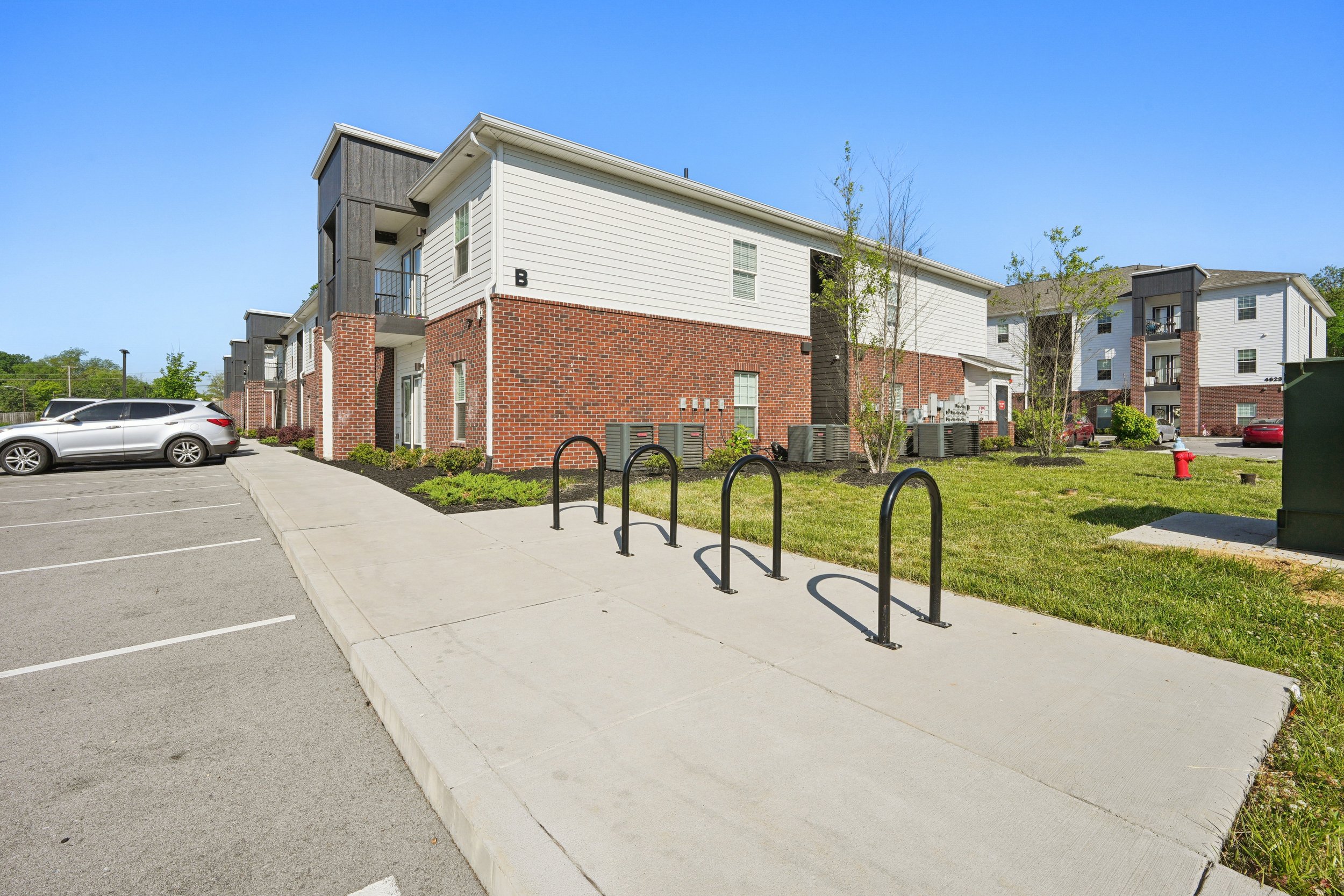 Apartment complex with white siding and brick lower walls, parking lot with silver car, paved sidewalk, small trees, green grass, and a fire hydrant under a clear blue sky.