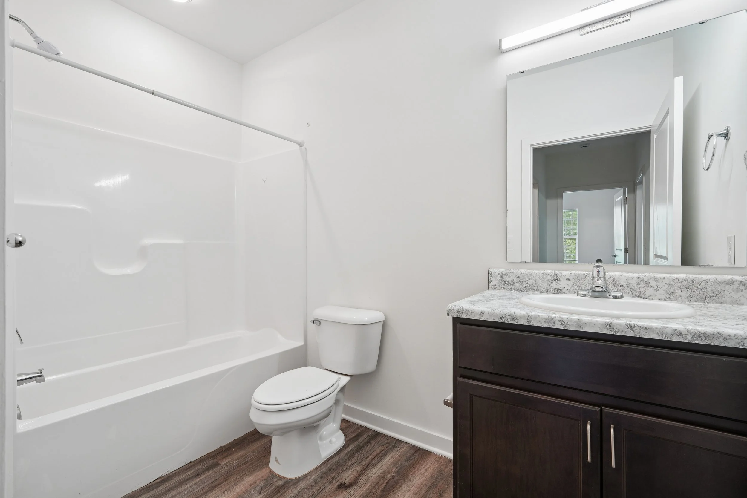 A modern bathroom with a white bathtub and shower, a toilet, a mirror above a marble countertop with a sink, and dark cabinetry. The walls are white, and there is wood-look flooring.