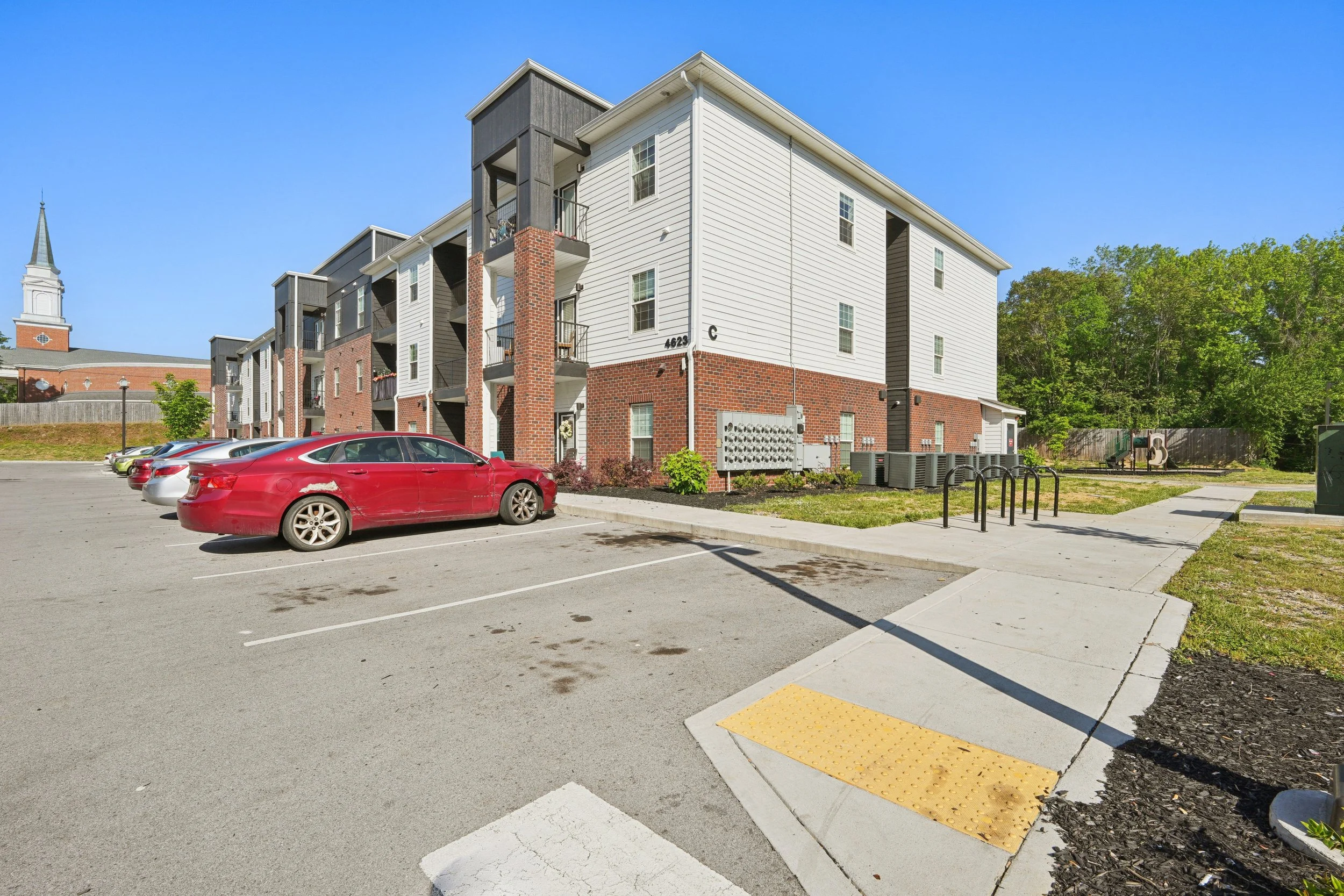 A parking lot with several parked cars in front of a multi-story residential building with white siding and brick accents. There is a church with a steeple in the background, green trees, and a clear blue sky.