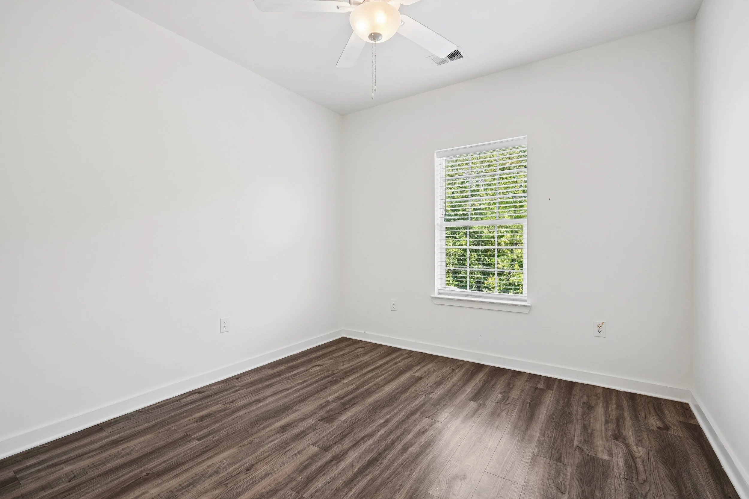 Empty room with white walls, a window with white blinds showing green trees outside, a ceiling fan with a light, and dark wood flooring.