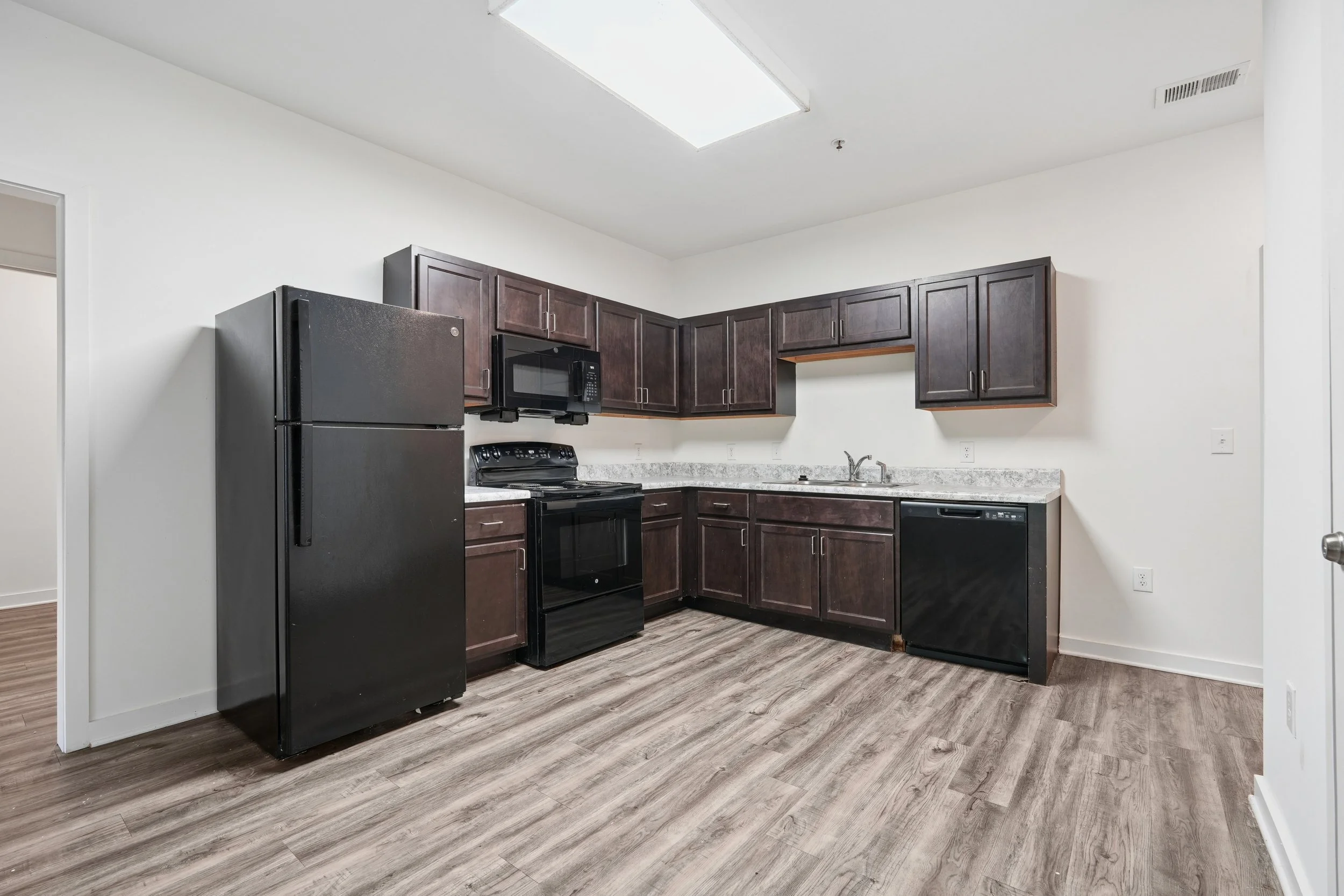 Kitchen with dark wood cabinets, black appliances including a refrigerator, microwave, and stove, white walls, and wood-look flooring.