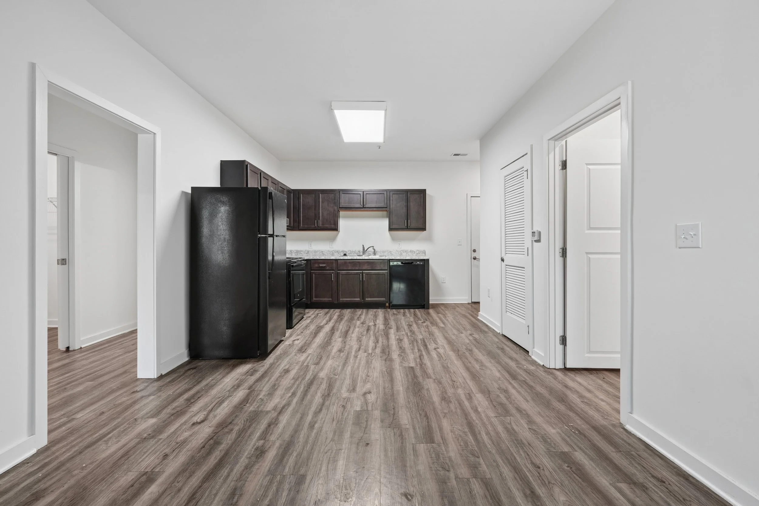 Empty kitchen with dark wooden cabinets, black refrigerator, and wood-look flooring.