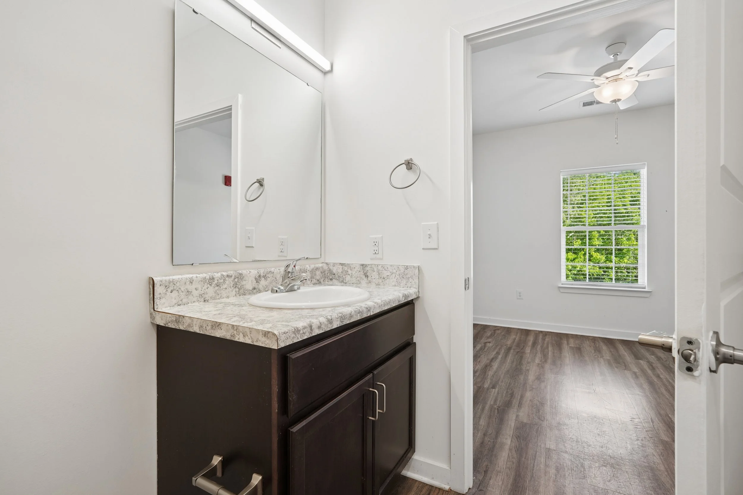 Bathroom with a mirror, sink with countertop, dark wooden vanity, and a view into a room with a ceiling fan and window.