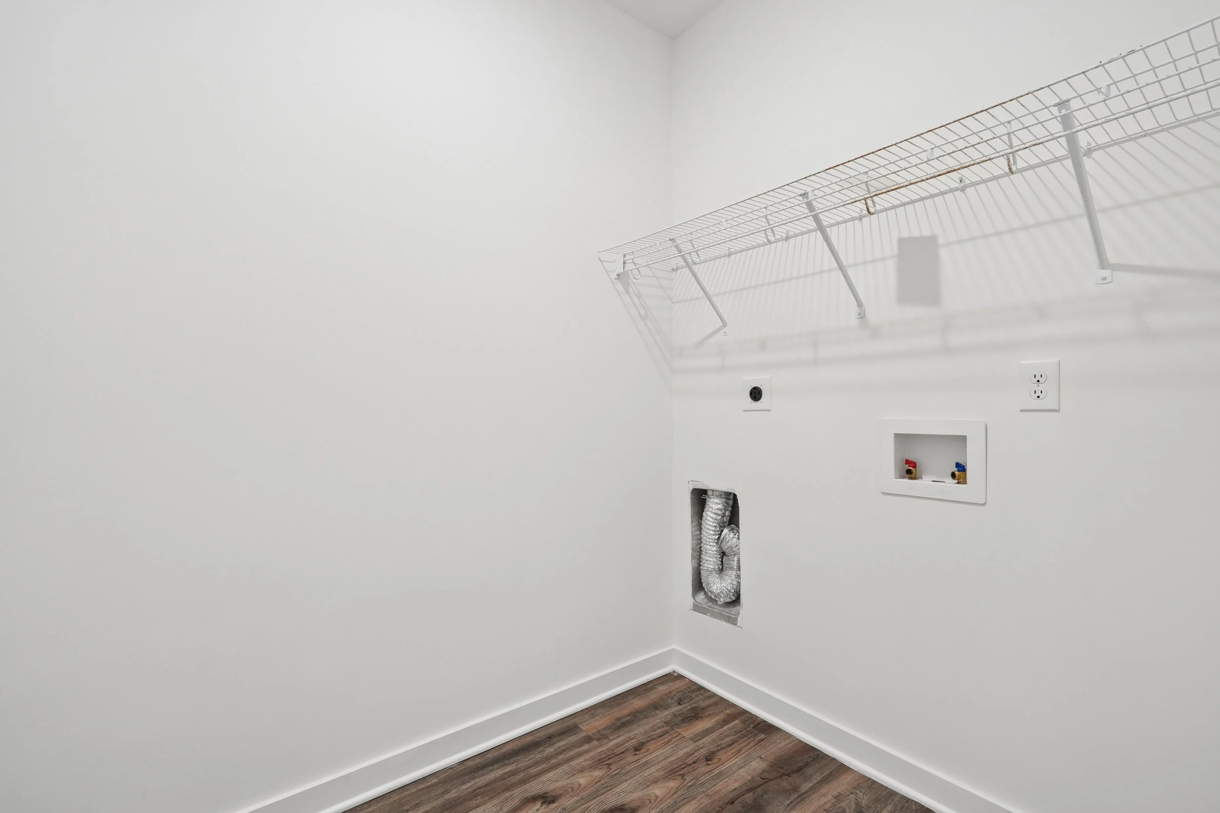 Empty laundry room with white walls, wire shelf, electrical outlets, and vent duct, with wooden flooring.