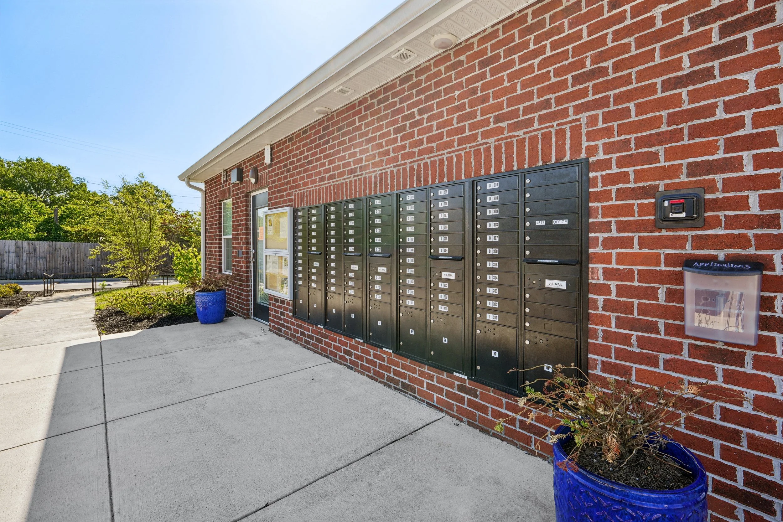A row of black metal mailboxes mounted on a red brick building exterior, with a blue planter containing a small plant and a sidewalk in front. Trees and a wooden fence are visible in the background under a clear blue sky.