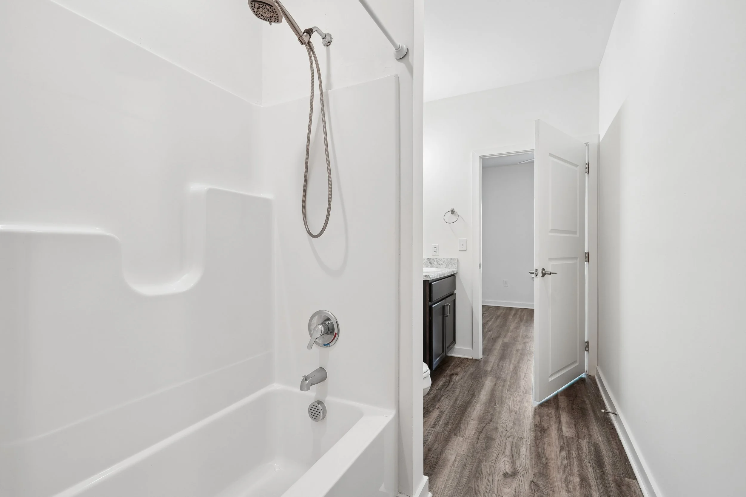 An image showing the interior of a bathroom with a walk-in shower and a view into an adjoining room with a dark cabinet and a granite countertop.