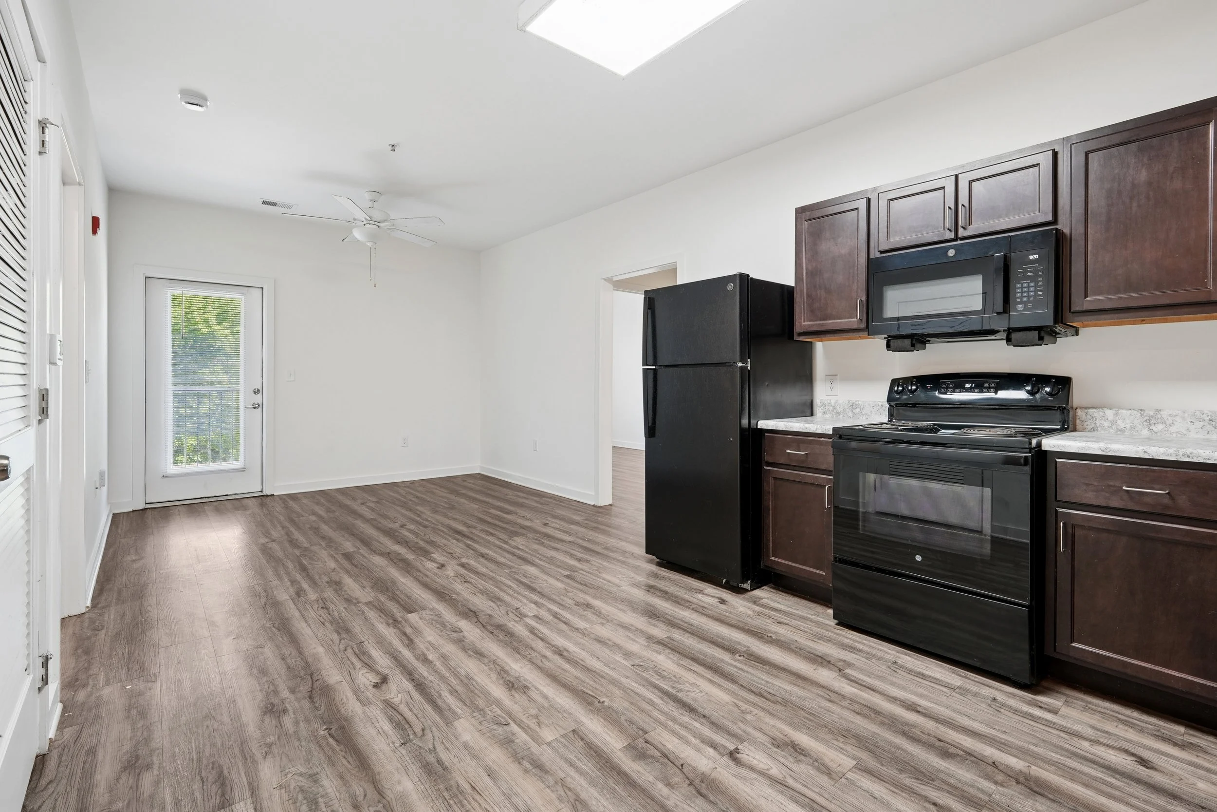 A kitchen with dark wood cabinets, black appliances including a microwave and refrigerator, and wood laminate flooring. There is a doorway, a window with blinds, and a ceiling fan. The room is well-lit with natural light.