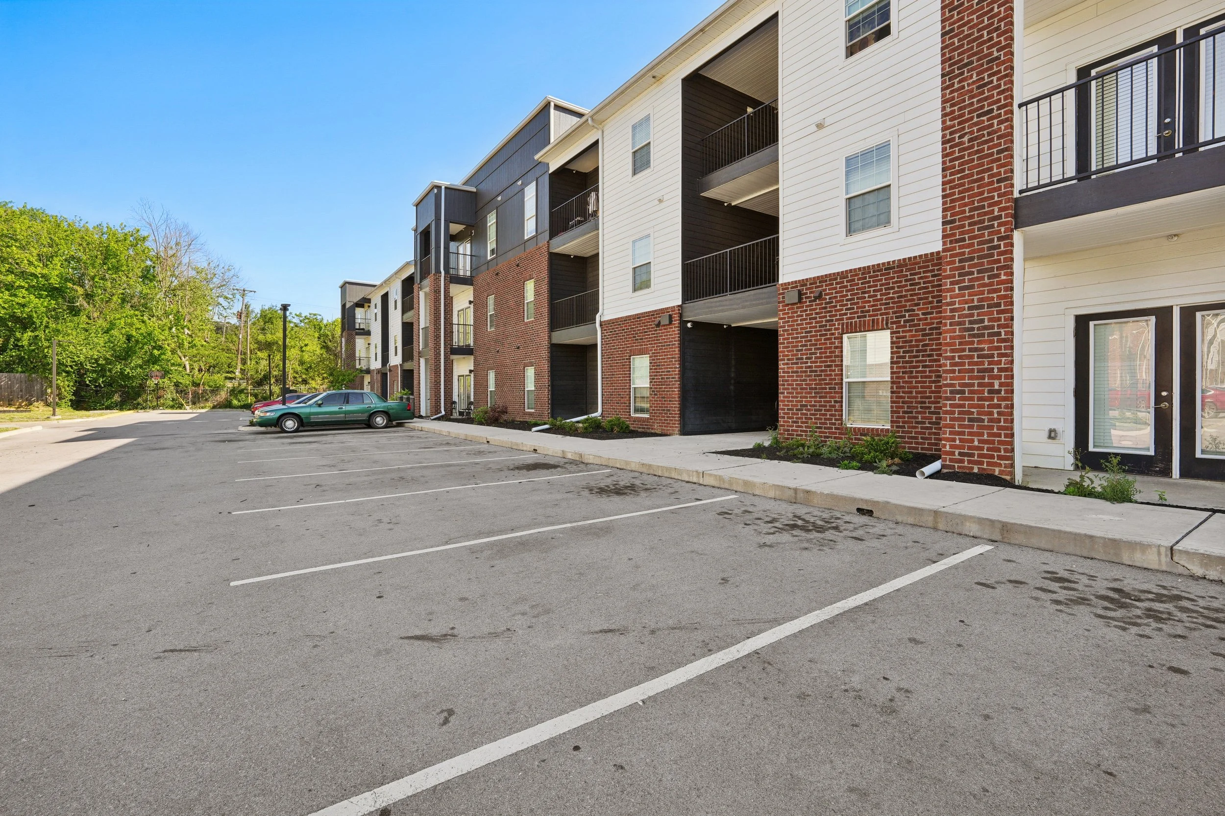 Empty parking lot in front of a multi-story residential building with cars parked along the curb, surrounded by greenery and a clear blue sky.