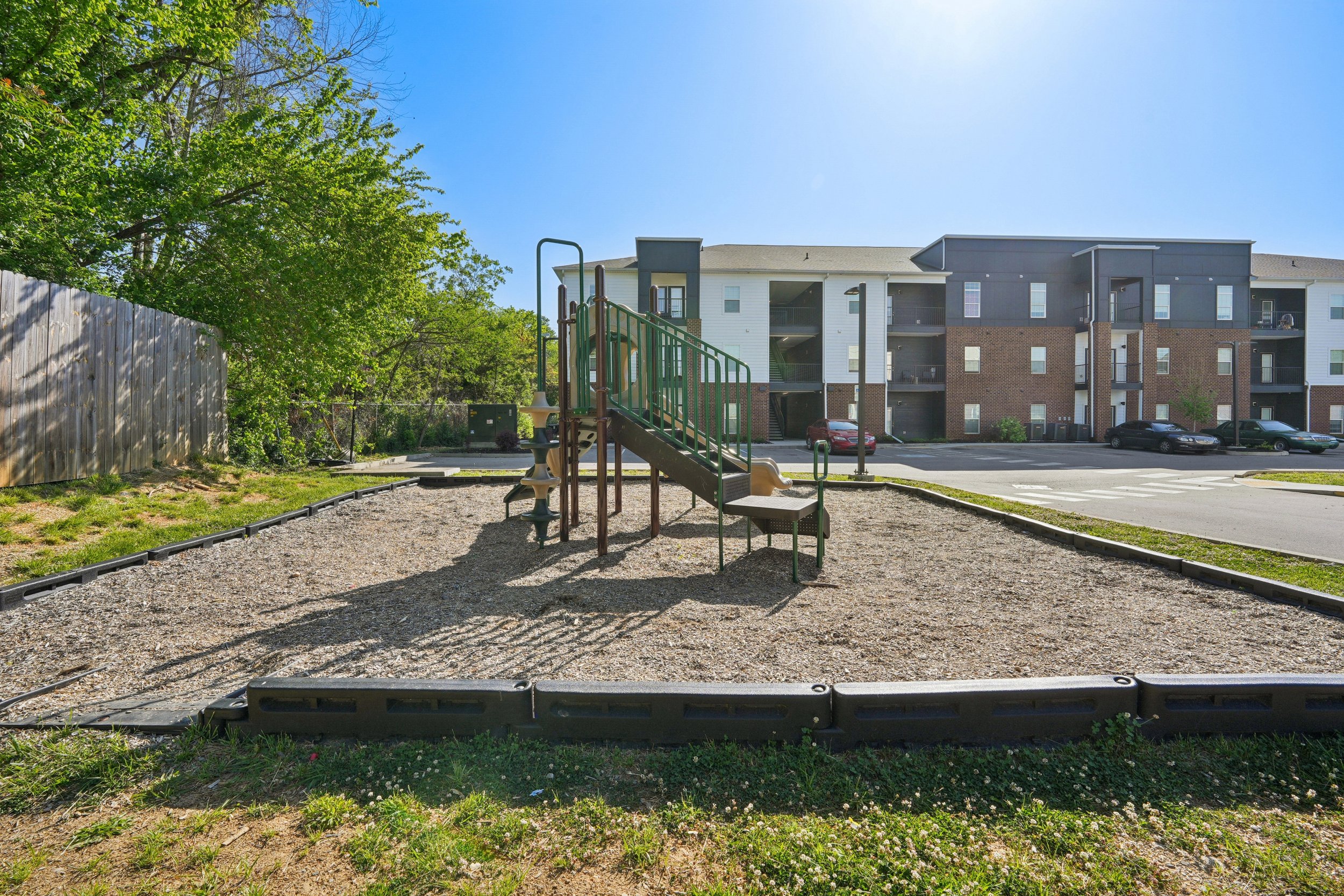 Playground with slide and climbing structure in front of a multi-story apartment complex, with a parking lot and cars, and a fenced yard with trees on a sunny day.