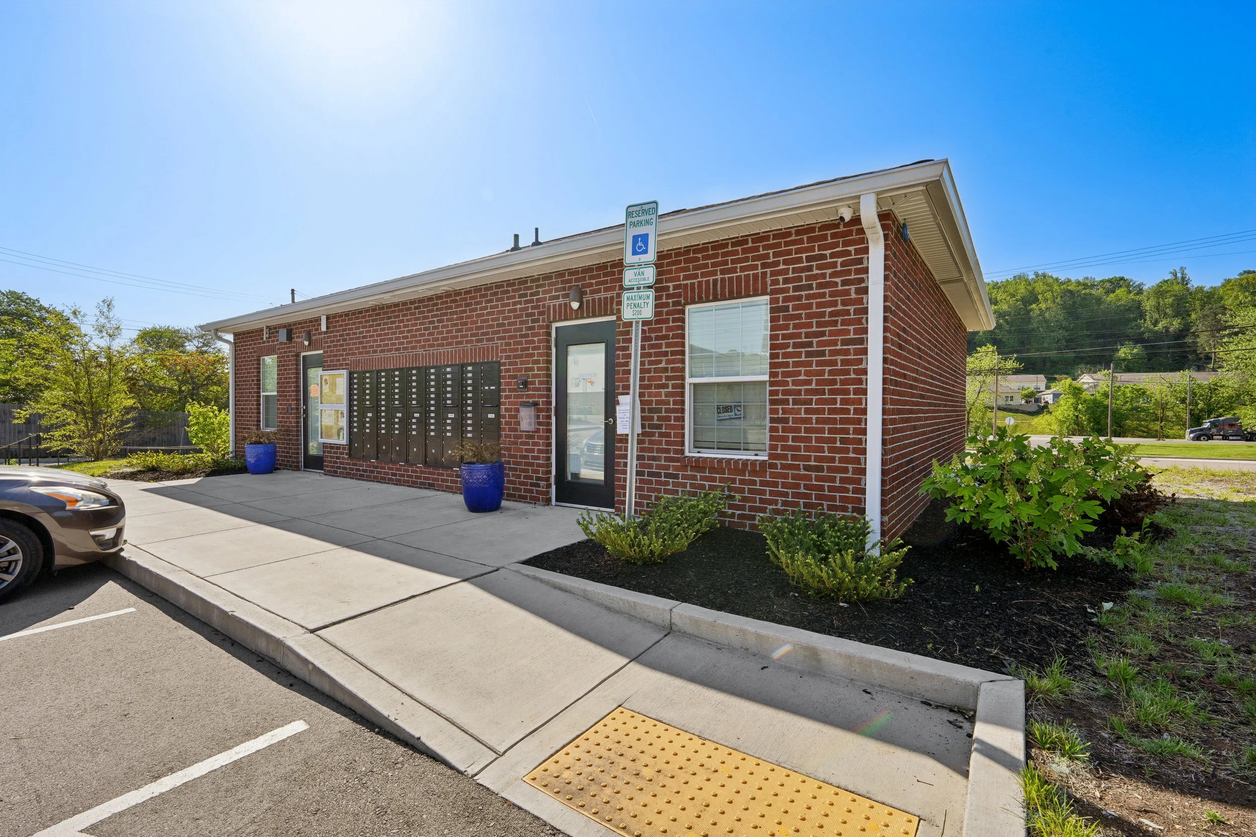 A brick building with mailboxes outside, designated handicapped parking sign, and sidewalk with curb ramp and yellow tactile paving, surrounded by greenery and blue sky.