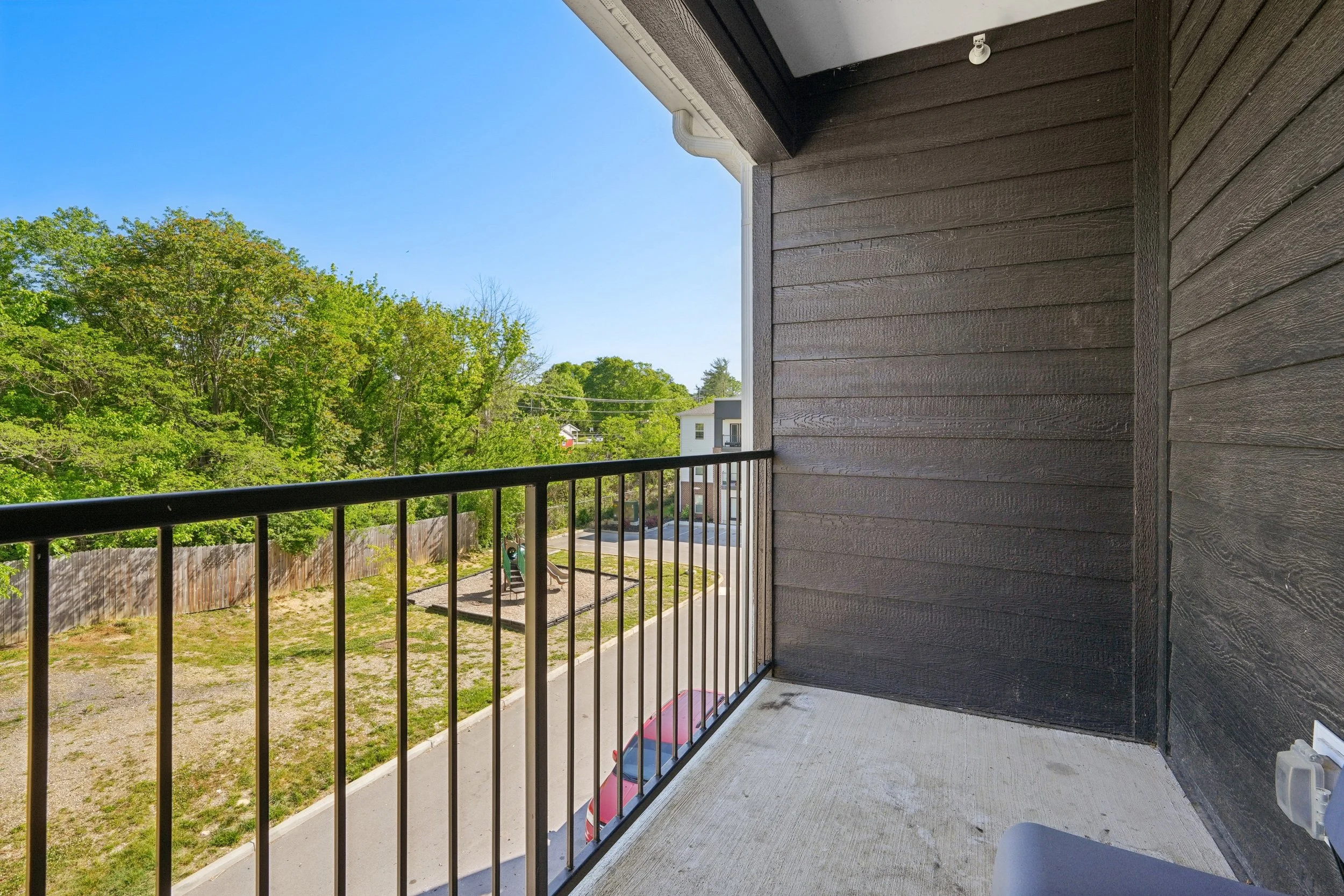 View from a balcony showing a black metal railing, wooden side wall, and the outdoors with a yard, trees, and houses under a clear blue sky.