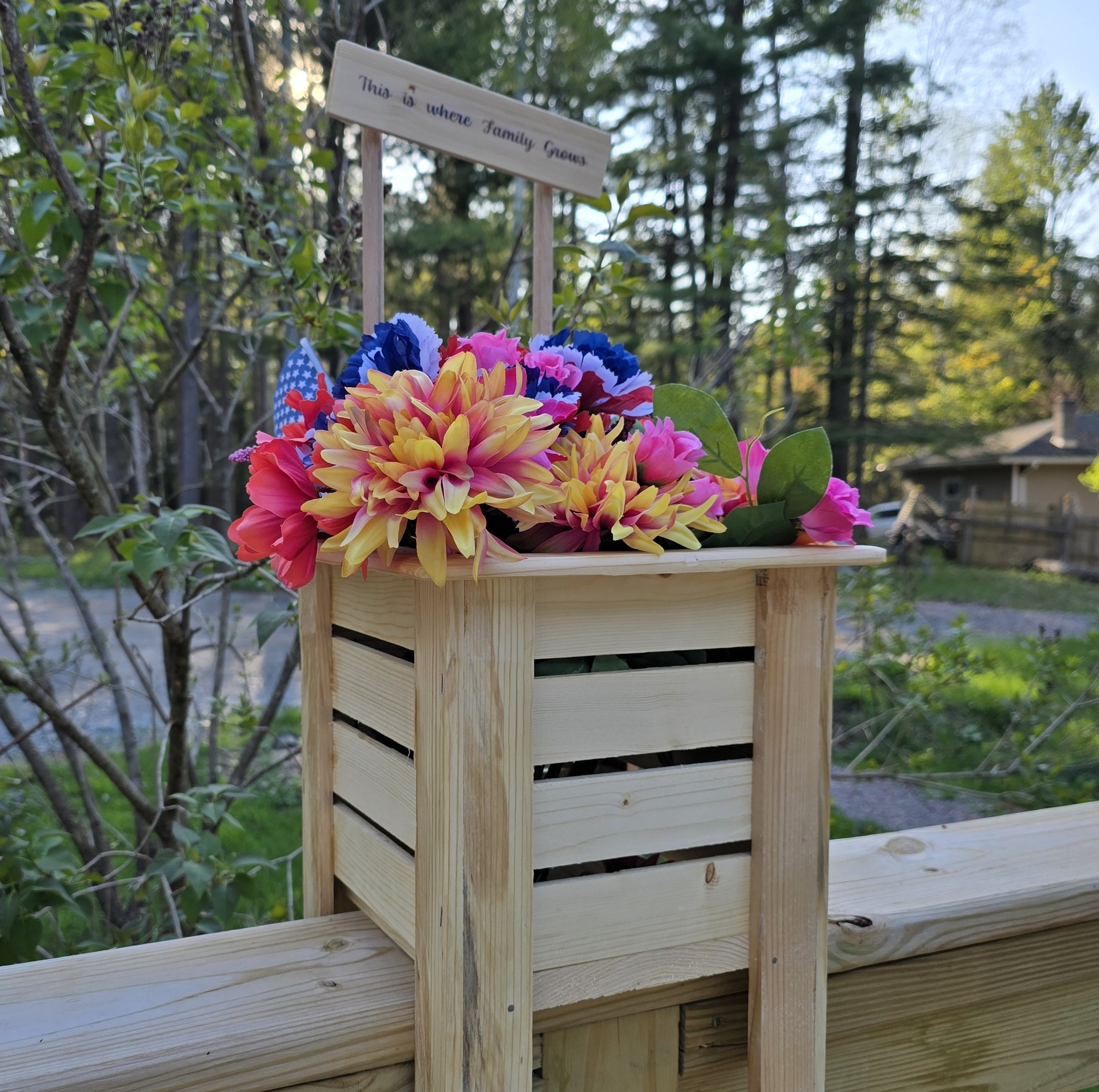 A wooden memorial stand with artificial flowers in red, yellow, pink, blue, and white, along with small American flags, sitting outdoors with trees and a house in the background.