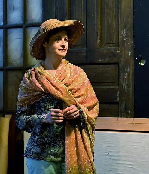 A woman wearing a large straw hat and a multicolored shawl, standing indoors in front of a wooden wall and window, holding a small black object.