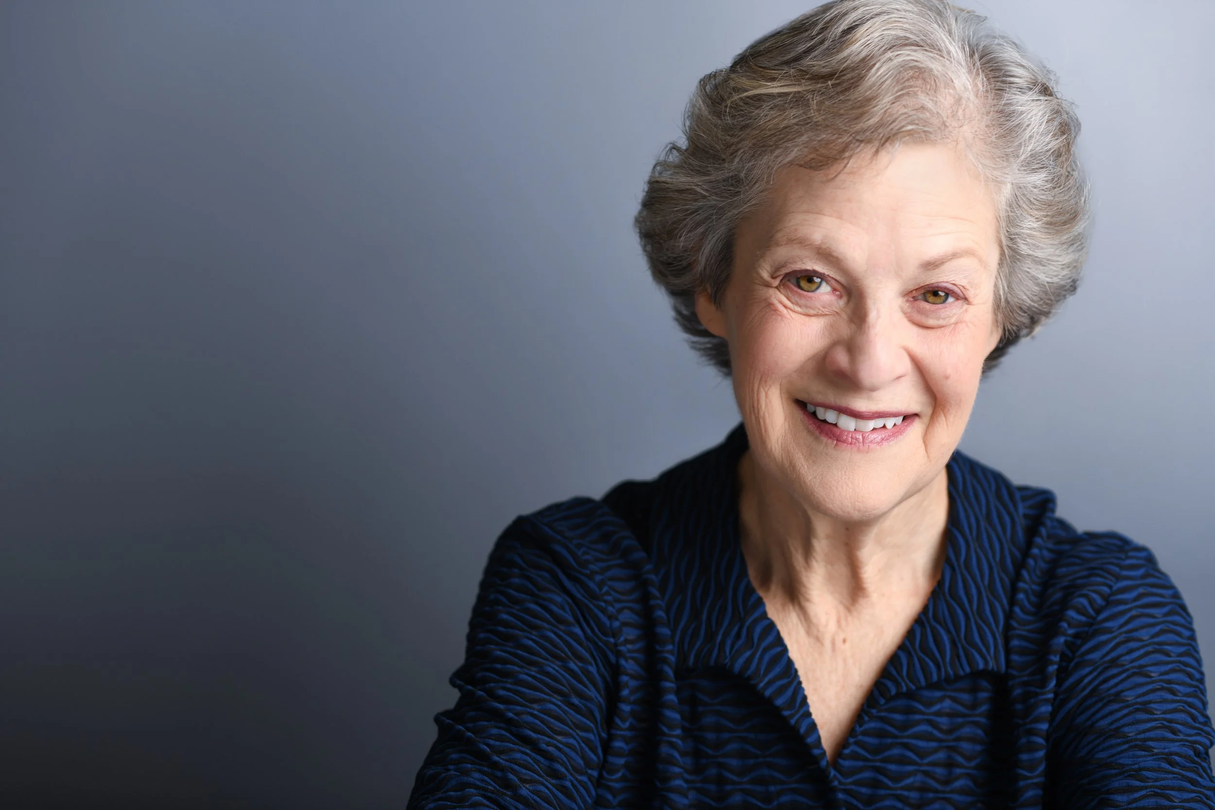 Close-up of a woman with gray hair smiling, wearing a dark blue textured top against a plain gray background.