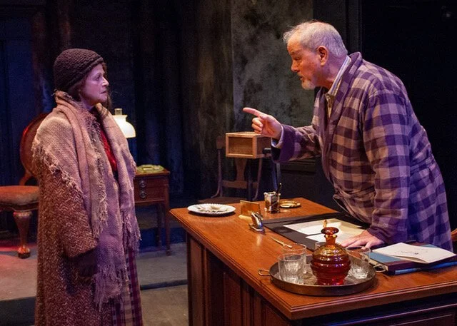Two actors on stage in a theatrical play. The woman on the left is wearing a long coat, a scarf, and a knitted hat. The man on the right is leaning on a wooden desk, pointing his finger, and wearing a plaid shirt and jacket. The scene appears to be set in a vintage or rustic room, with props including a tray, glass, and decorative items on the desk.