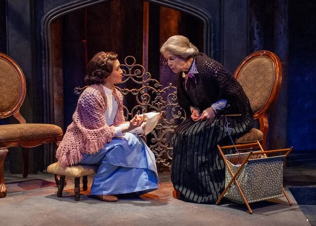 Two women, one young and one elderly, are engaged in a conversation on stage. The young woman is sitting on a footstool, holding a book, while the elderly woman is standing and leaning forward. The setting features a fireplace and ornate chairs.
