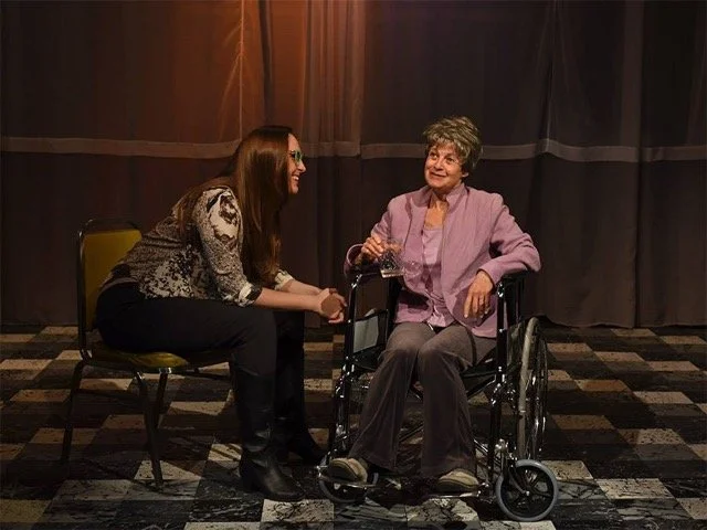 Two women are sitting together in a dimly lit room with curtains in the background. One is sitting on a chair, leaning forward, smiling and holding the woman's hand, who is sitting in a wheelchair. The woman in the wheelchair is smiling, holding a glass of water, and wearing a purple jacket.