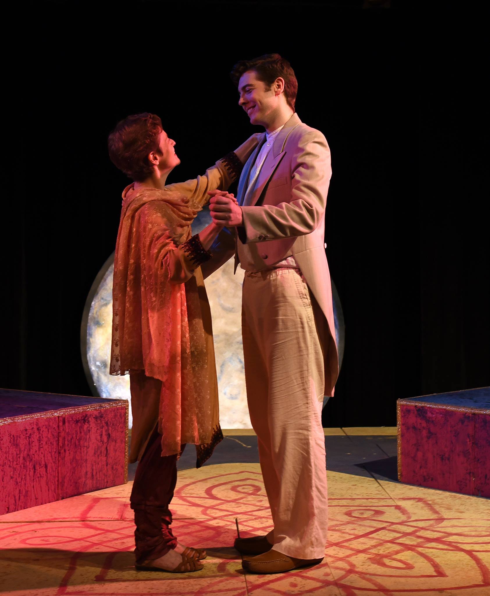 A young boy and a man are standing on a stage, holding hands, and looking at each other. The boy wears a brown jacket with embroidery and 'cuffs' at the sleeves, while the man wears a light-colored suit. The background includes a full moon, dark sky, and stage set pieces.