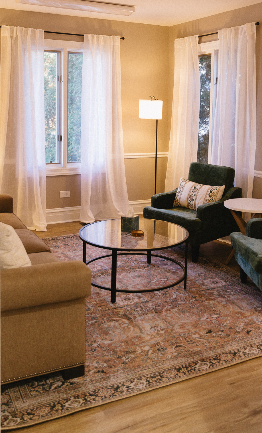 A cozy living room with a beige sofa, green armchairs, a round glass coffee table, a standing lamp, hardwood floors, and large windows with white curtains, allowing natural light to enter.