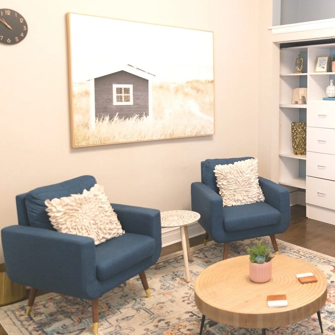 Living room with two blue armchairs with white textured pillows, a round wooden coffee table with a potted plant, a patterned rug, a white bookshelf with decorative items, and a wall art depicting a small dark house in a field.
