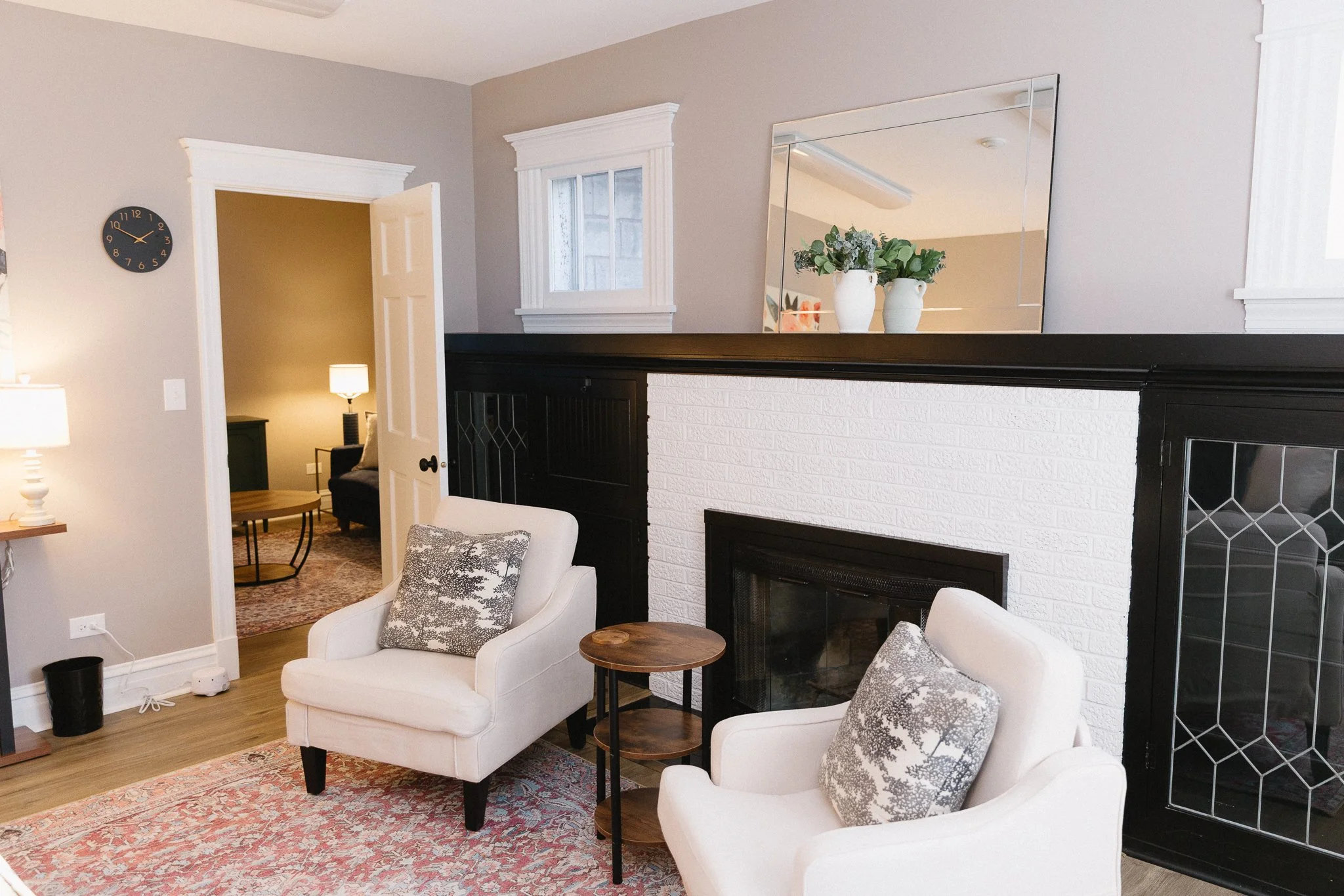 Living room with white armchairs, side tables, a fireplace, mirror, and houseplants on mantel, with a doorway to a lit room in the background.