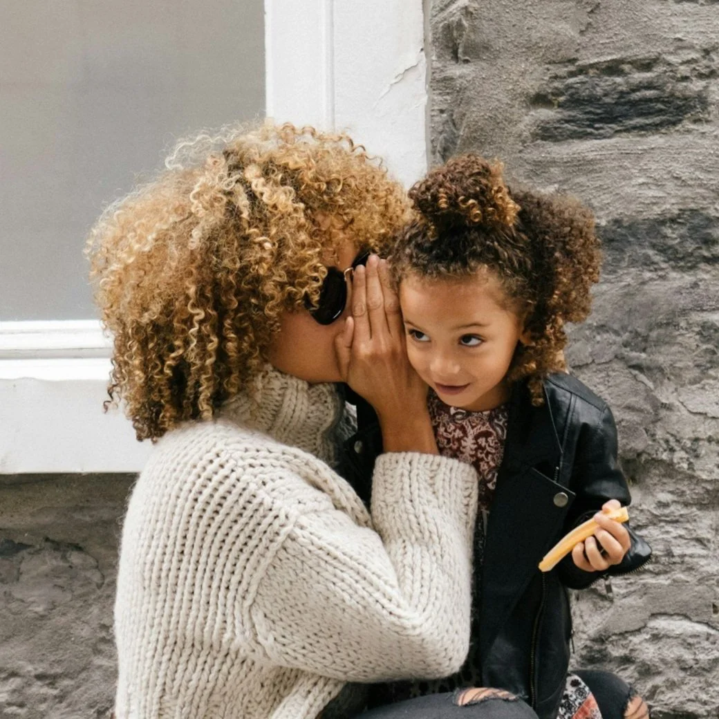 A woman whispering into a young girl's ear outside against a stone wall.
