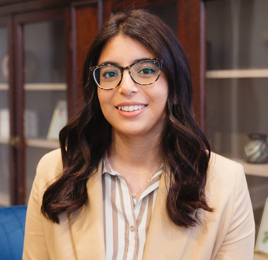A woman with long dark wavy hair, wearing glasses, a beige blazer, and a striped shirt, smiling at the camera in an indoor setting with shelves in the background.