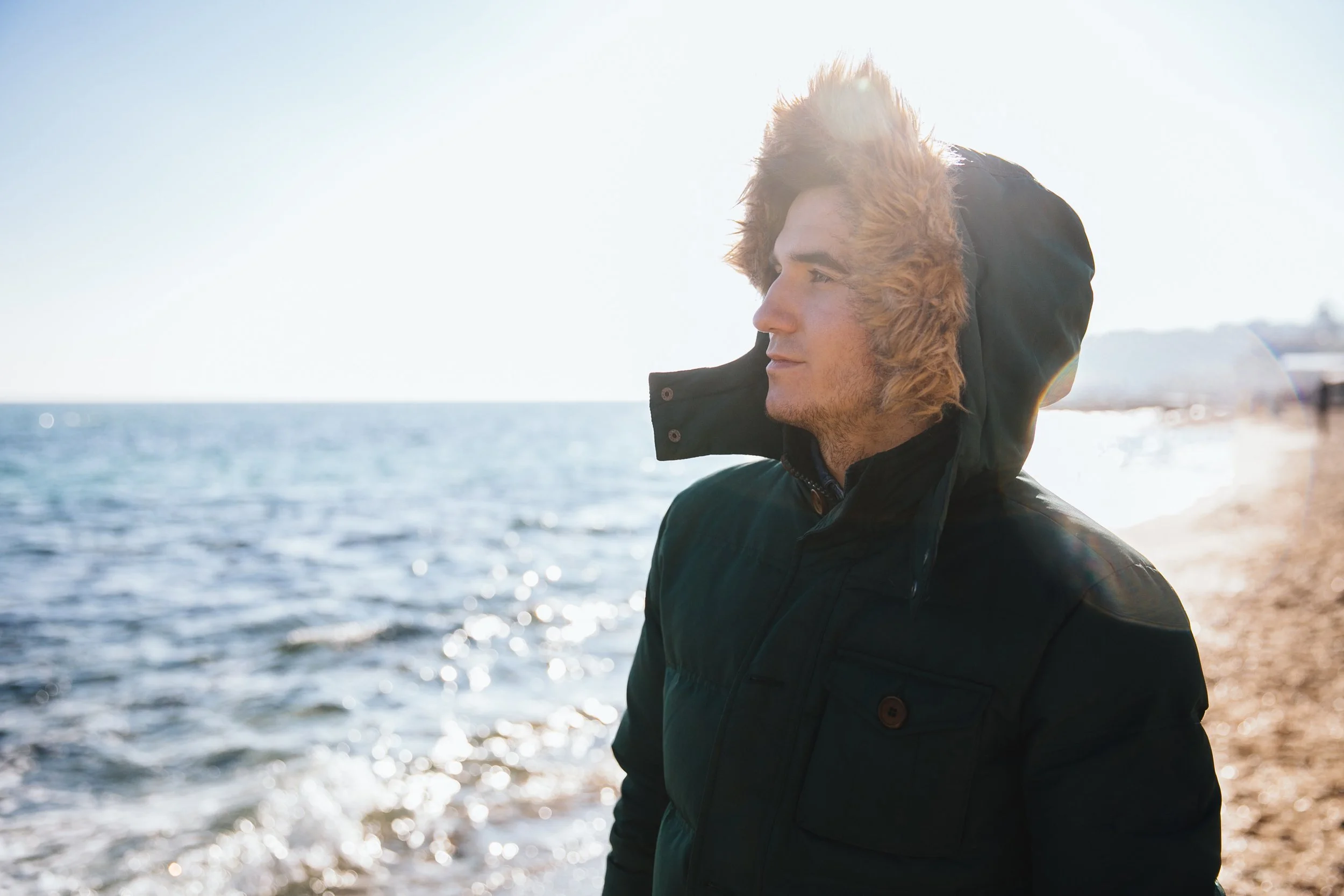 A young man with light skin and brown hair is standing on a beach, facing to the right, wearing a black winter coat with a hood with faux fur trim. The sun is shining brightly, and the ocean is in the background with gentle waves.