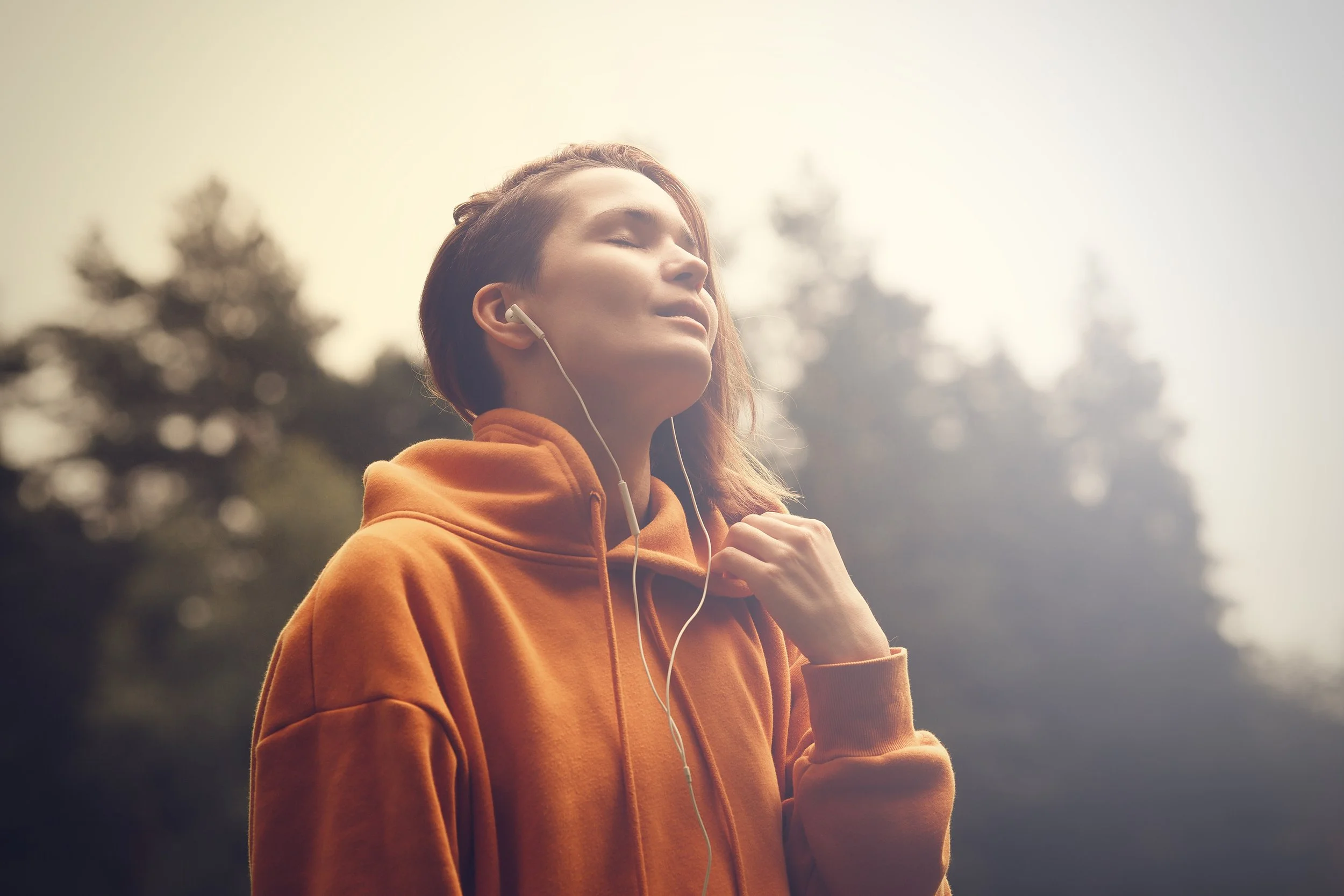 Young woman in orange hoodie enjoying listening to music with eyes closed in outdoor setting.