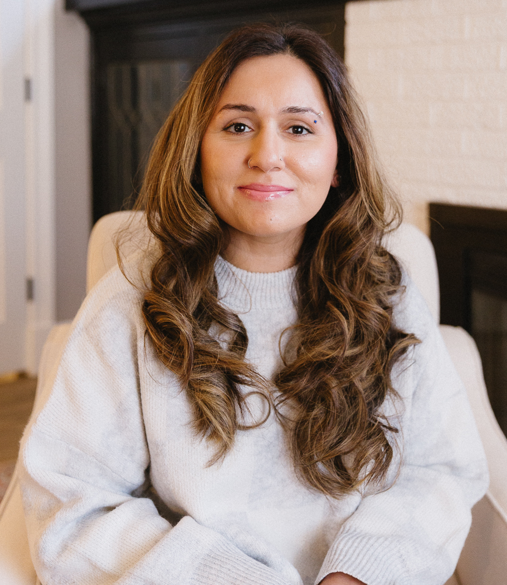 A woman with long, wavy brown hair wearing a gray sweater, sitting on a beige chair, smiling at the camera in a cozy room with a white brick wall and a dark piece of furniture in the background.