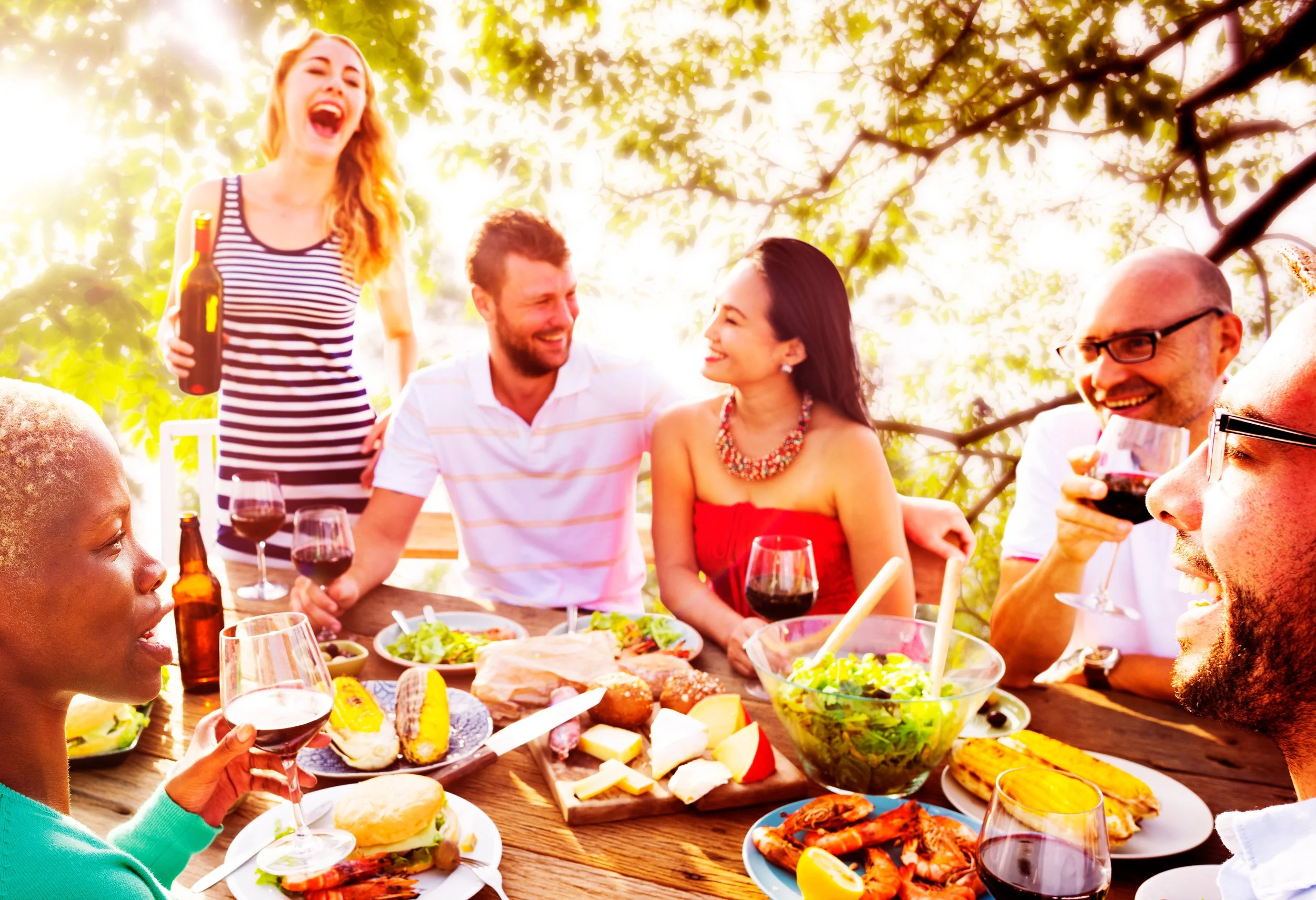 Group of people enjoying outdoor meal with food and drinks, surrounded by trees and sunlight.