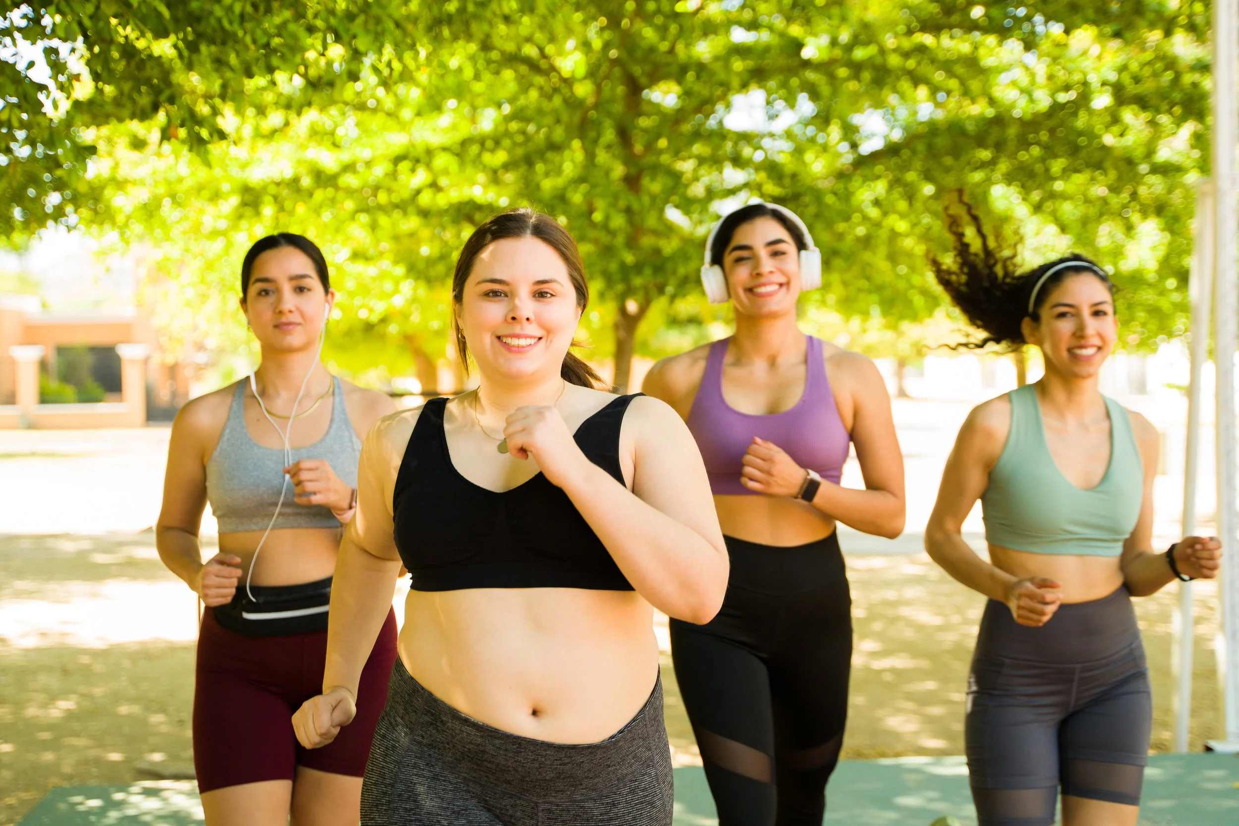 Four women jogging outdoors in a park on a sunny day, wearing athletic clothing and headbands. They are smiling and appear to be enjoying themselves.