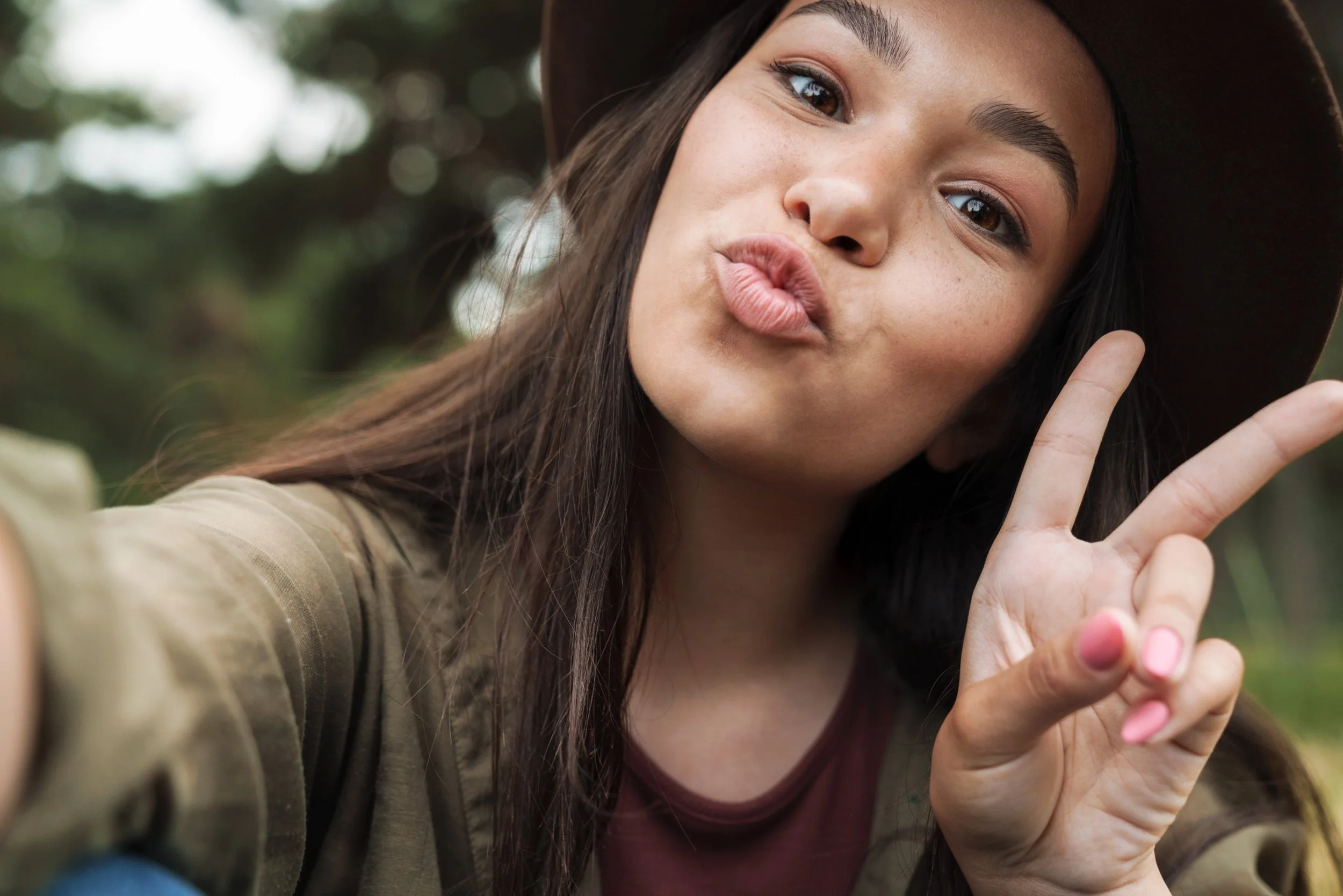 Close-up of a young woman with long dark hair making a kissing face, holding up her hand with two fingers in a peace sign, outdoors with a blurred background.