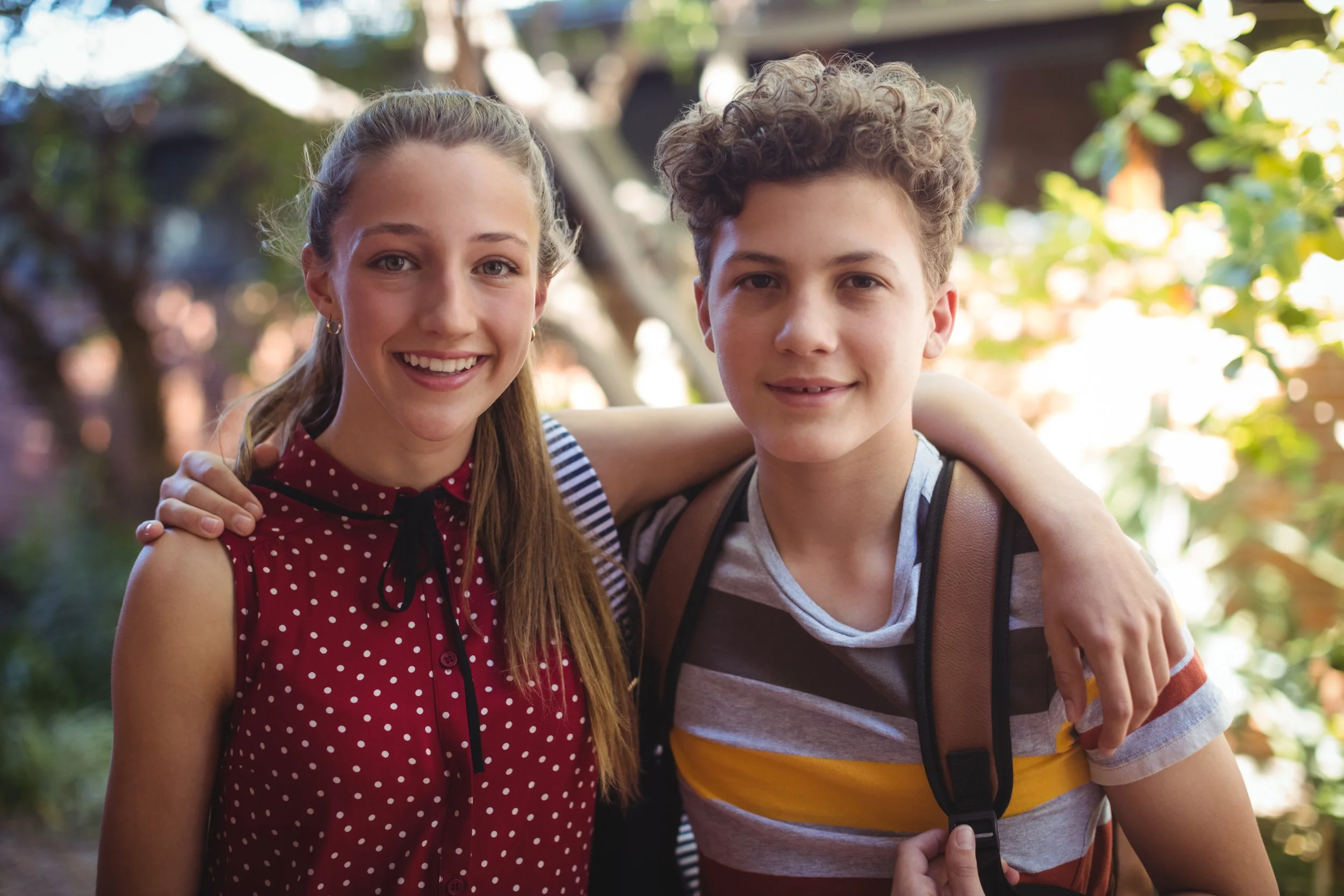 A smiling girl with brown hair in a ponytail and a smiling boy with curly hair, standing closely outdoors with arms around each other, in a park or garden area with green foliage.